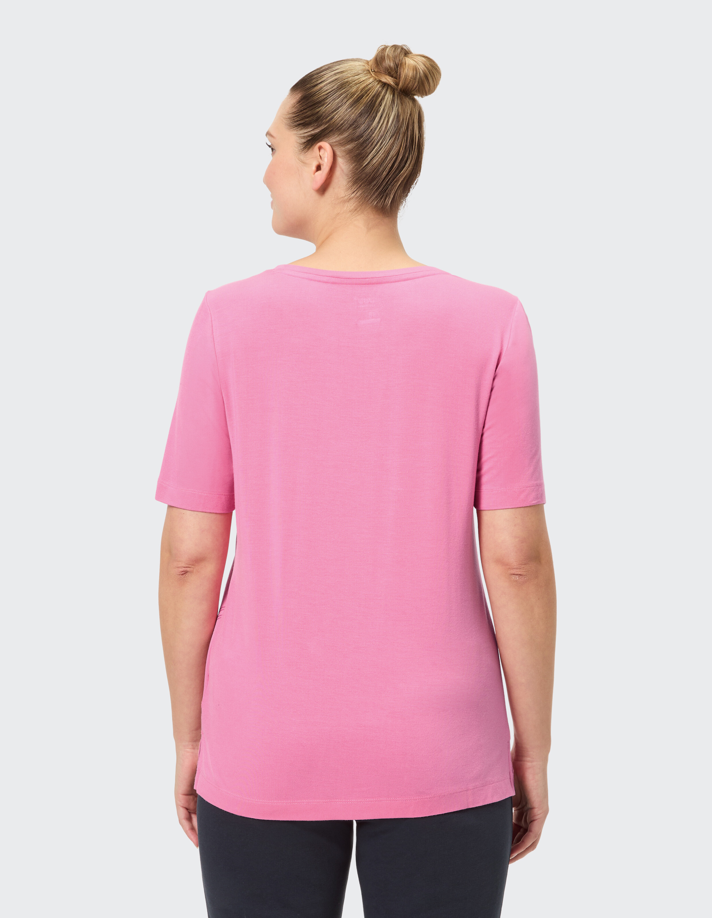 Person von hinten in pinkem T-Shirt mit hochgestecktem Haar, schwarzer Hose, vor hellem Hintergrund.