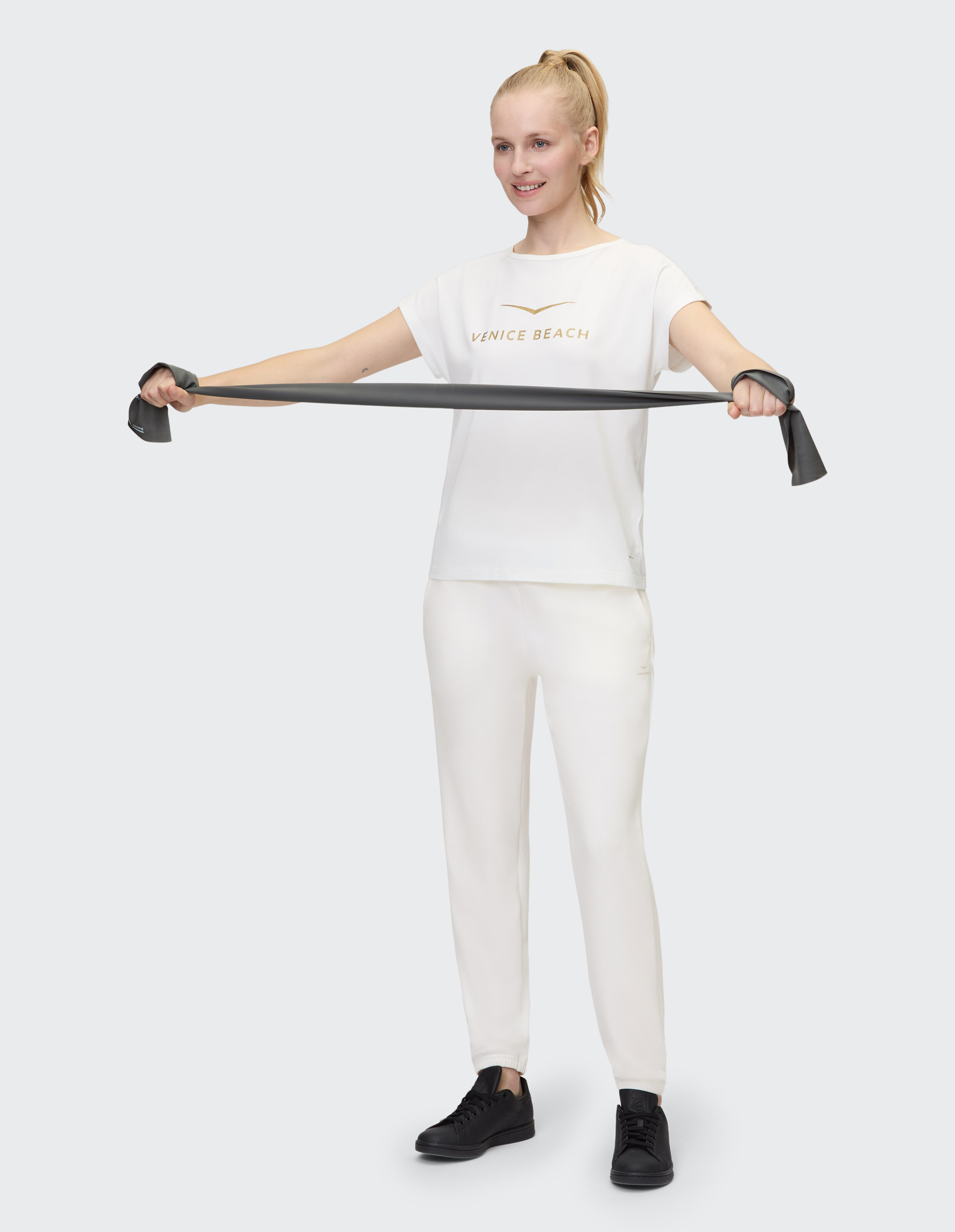 Frau in weißer Jogginghose und T-Shirt von Venice Beach trainiert mit einem Widerstandsband.