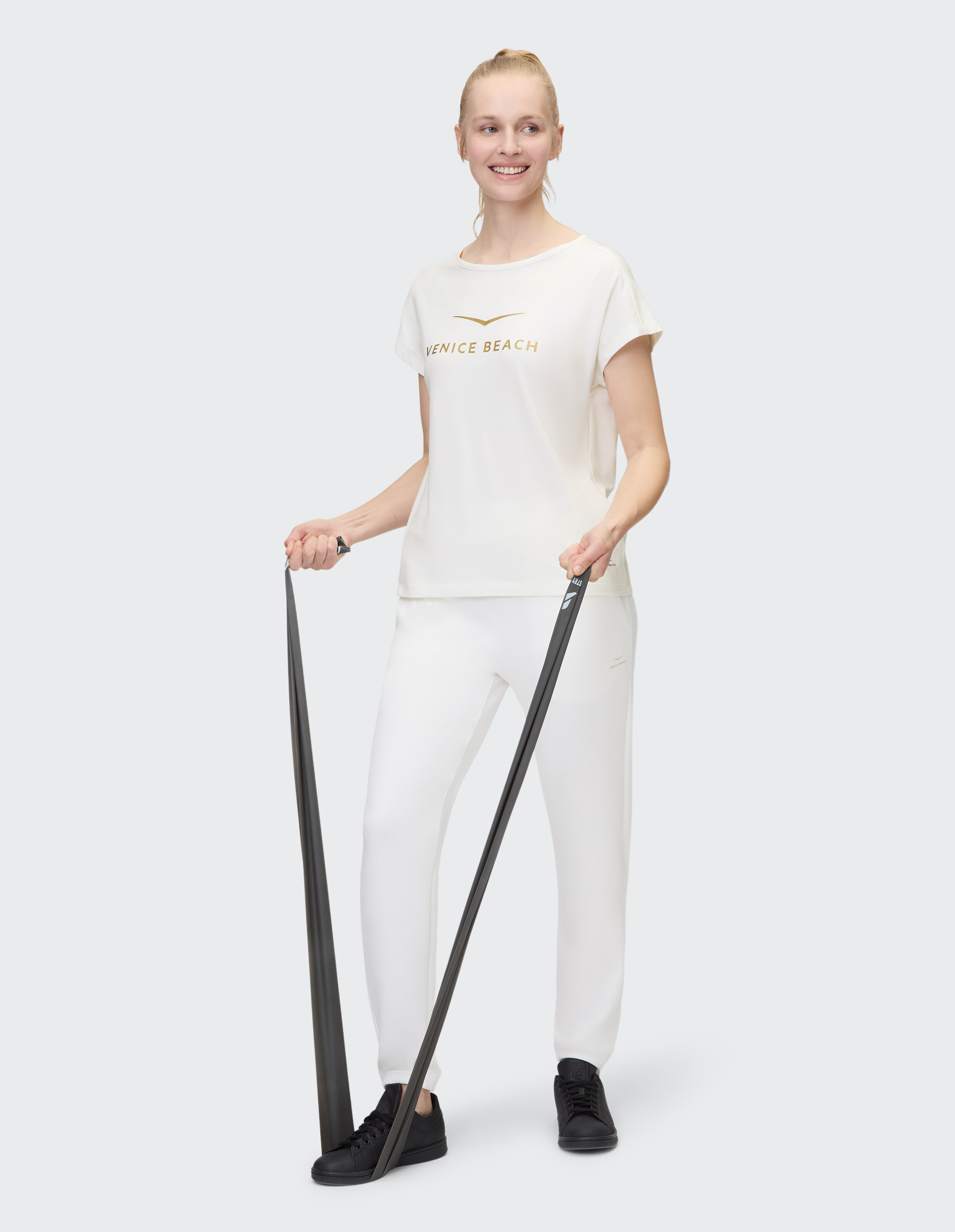 Frau in weißer Freizeitkleidung mit „Venice Beach“-Aufdruck, hält Widerstandsbänder in der Hand.