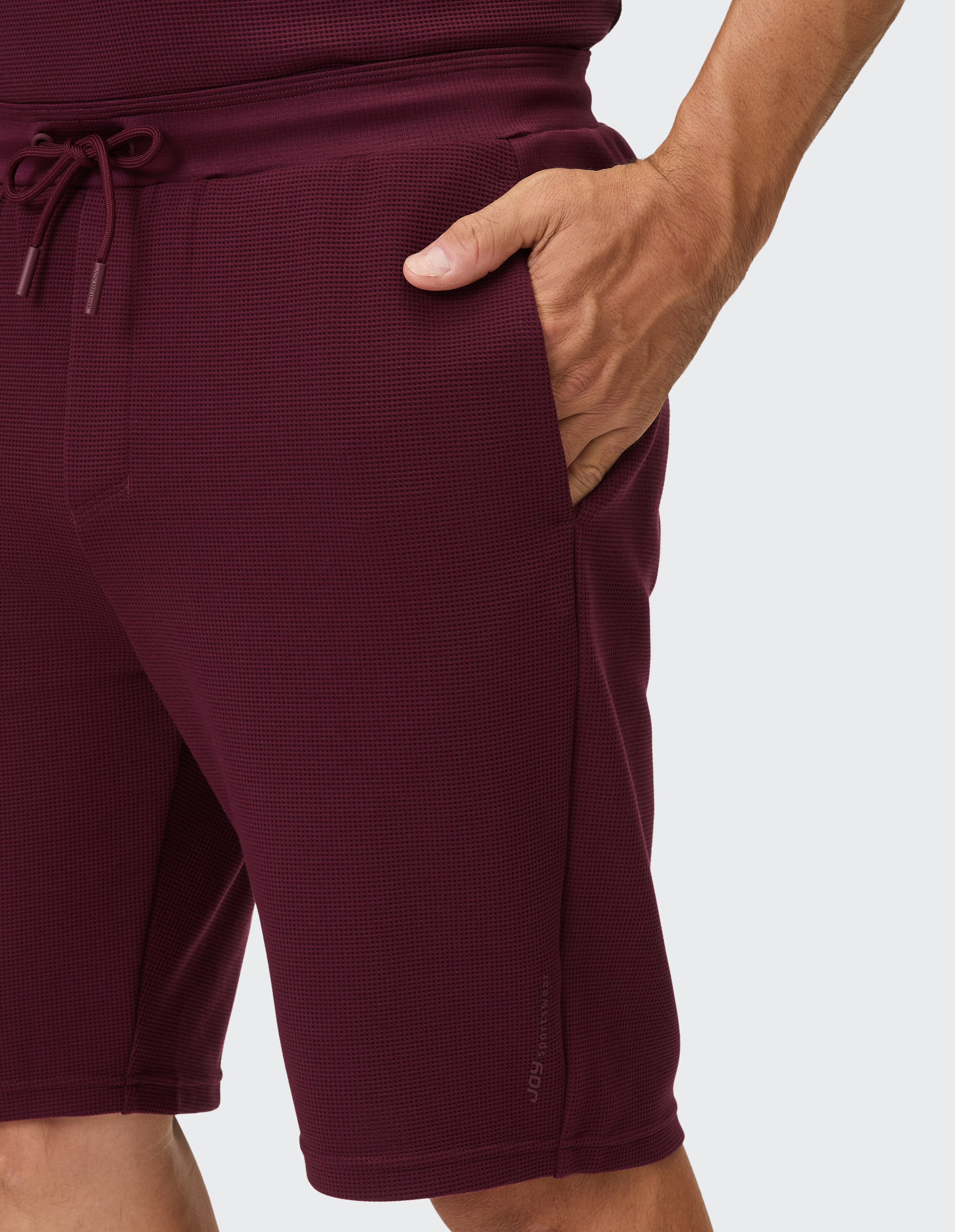 Eine weinrote Hose mit Waffelmuster und Kordelzug am Bund wird von einer Hand in der Tasche gehalten.