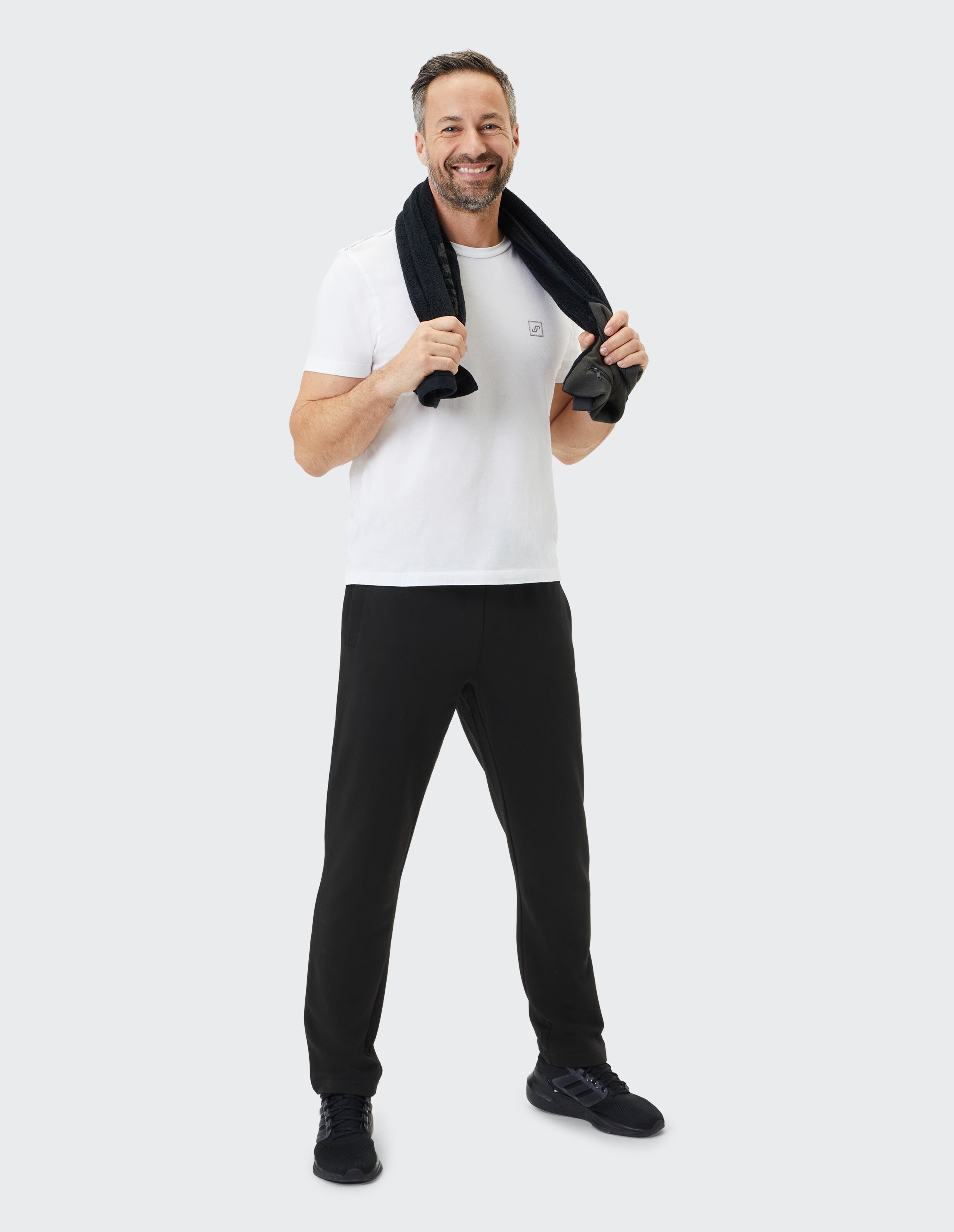 Mann in weißem T-Shirt und schwarzer Hose steht lächelnd mit schwarzem Handtuch über den Schultern vor grauem Hintergrund.