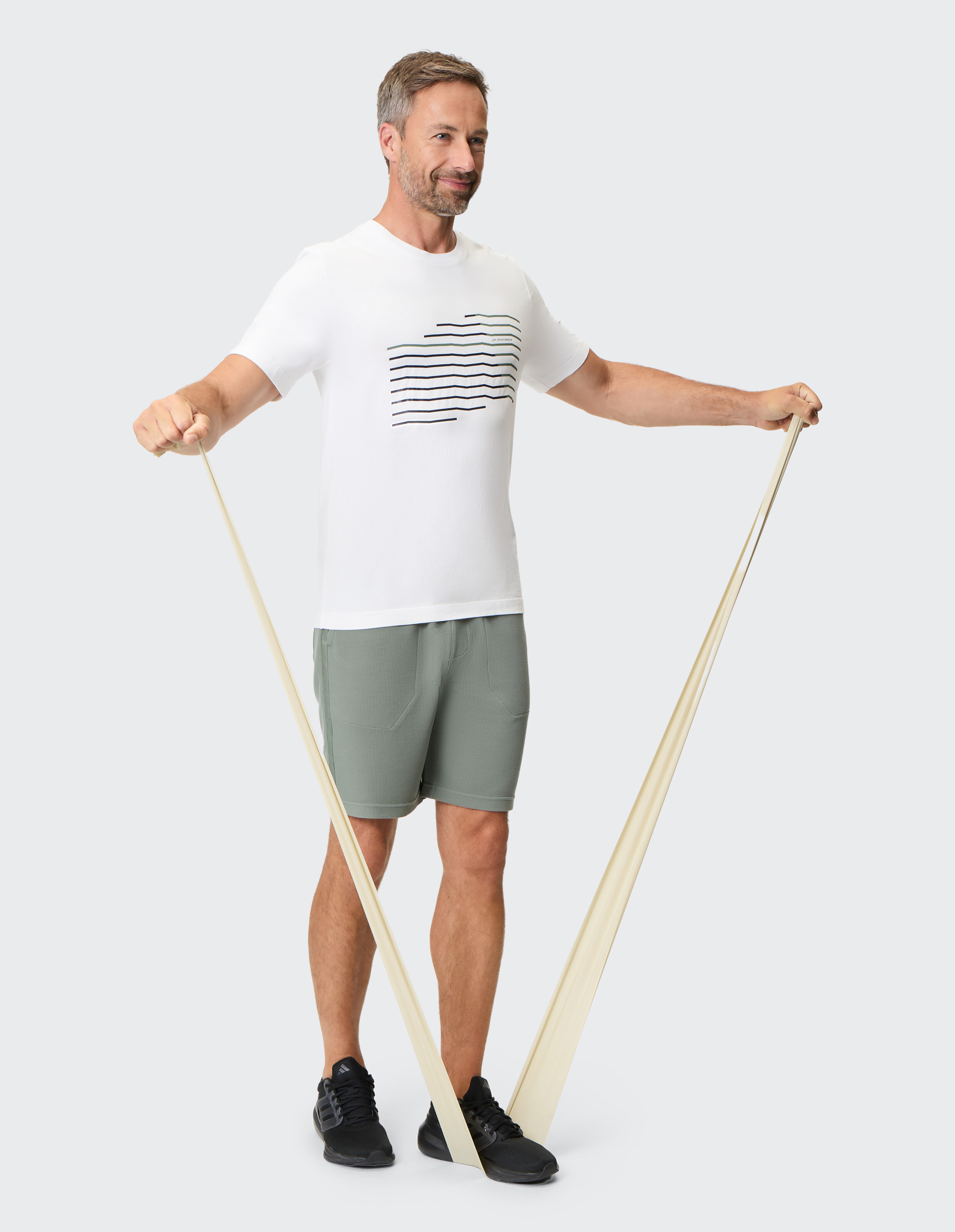 Ein Mann trainiert mit einem beigen Widerstandsband, trägt ein weißes T-Shirt und grüne Shorts, steht auf grauem Boden.