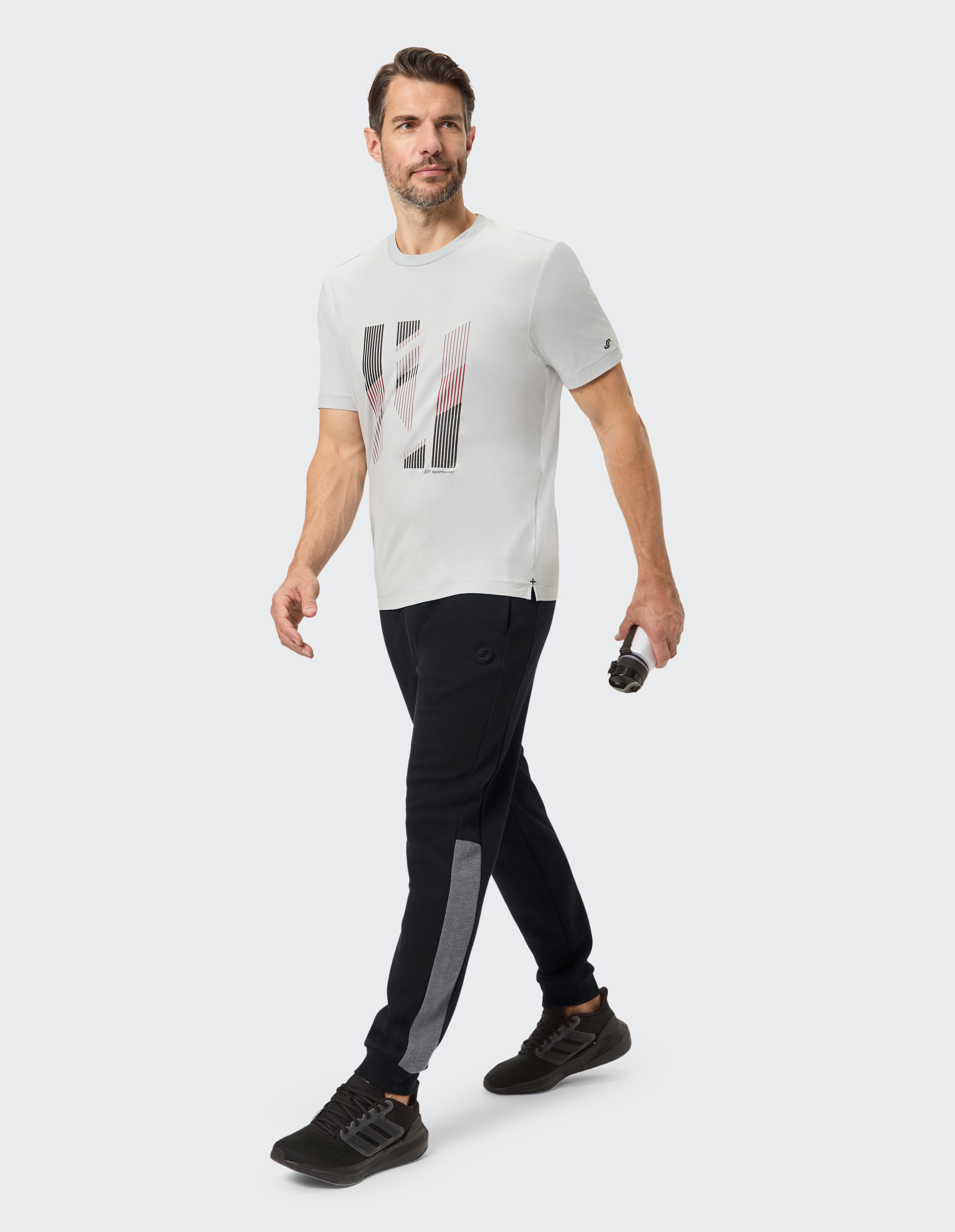 Mann in einem hellgrauen Grafik-T-Shirt und schwarzen Jogginghosen, der lässig geht.