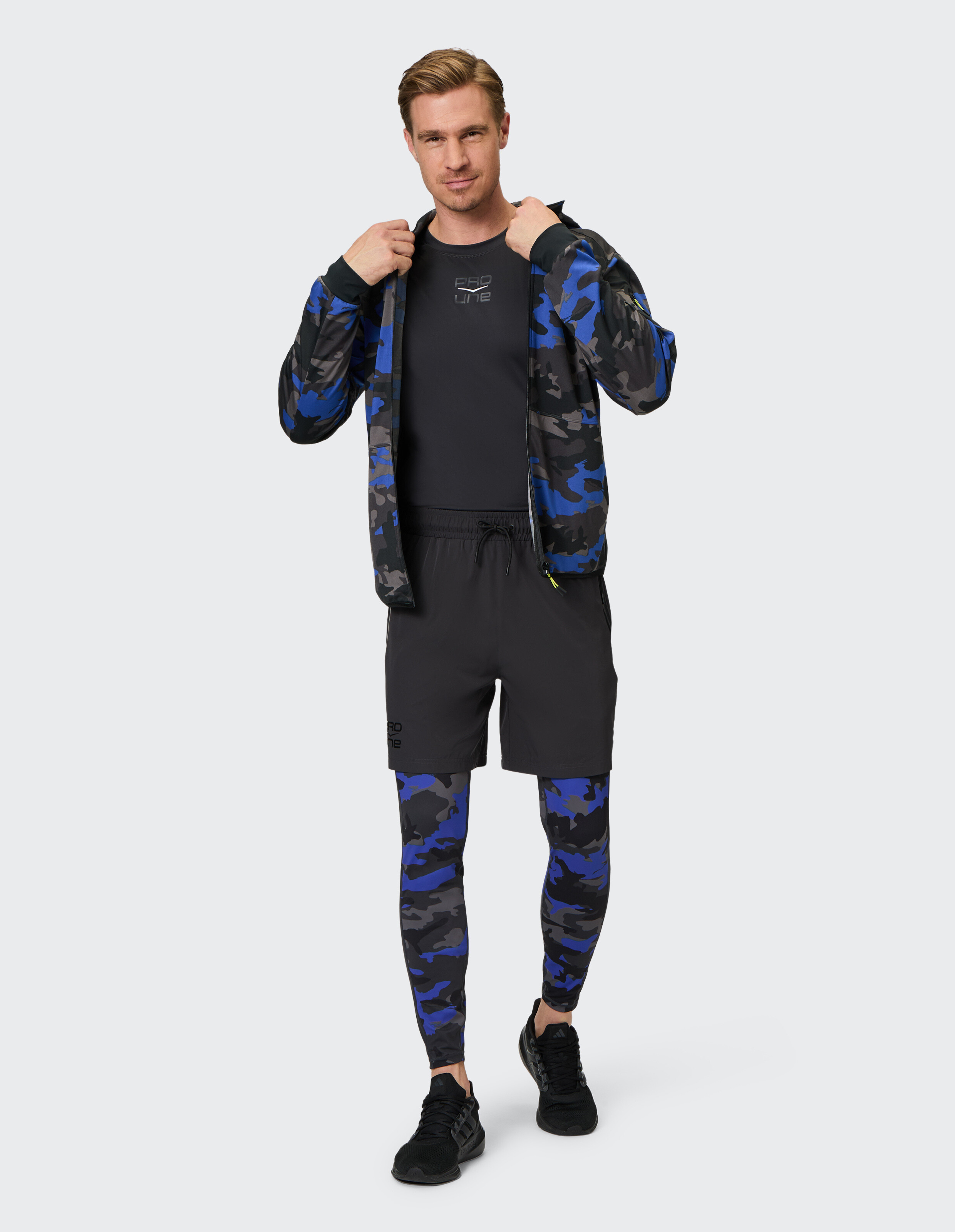 Mann trägt eine blaue Tarnjacke mit Reißverschluss, schwarze Shorts und passende Tarn-Leggings.