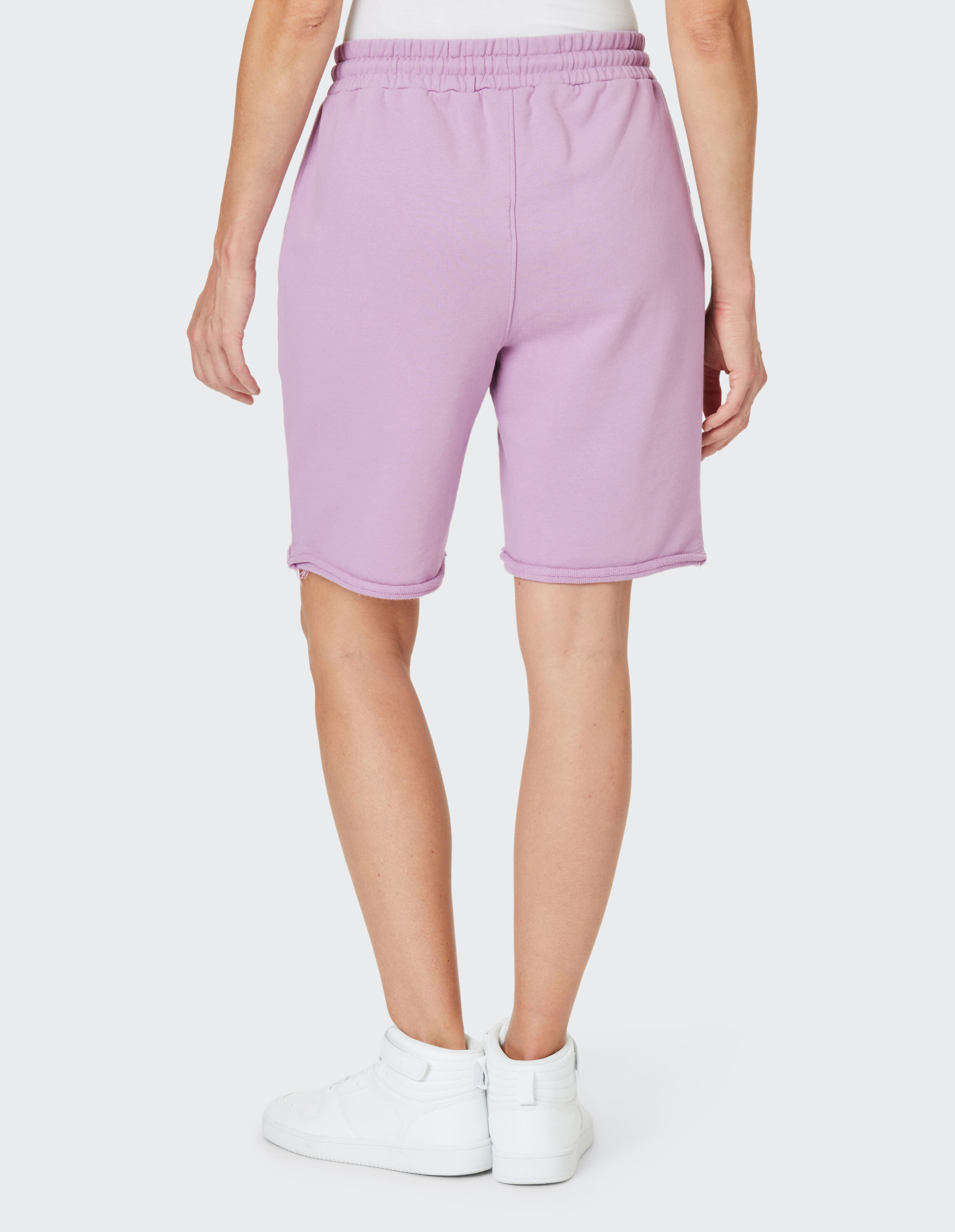 Person trägt lila Shorts und weiße Sneaker, steht mit dem Rücken zur Kamera auf hellem Hintergrund.