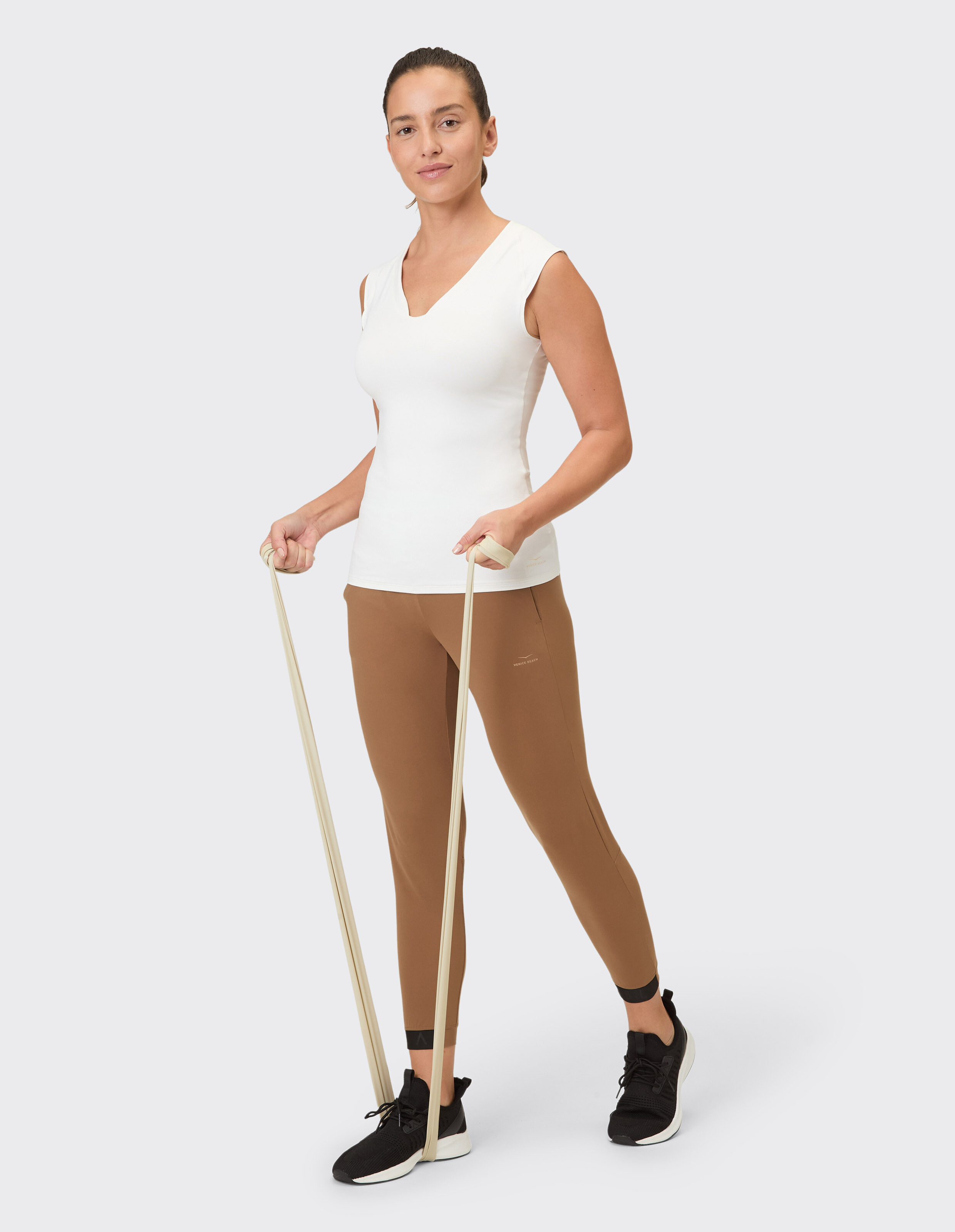 Frau in weißem Top mit V-Ausschnitt und braunen Leggings trainiert mit einem Widerstandsband.