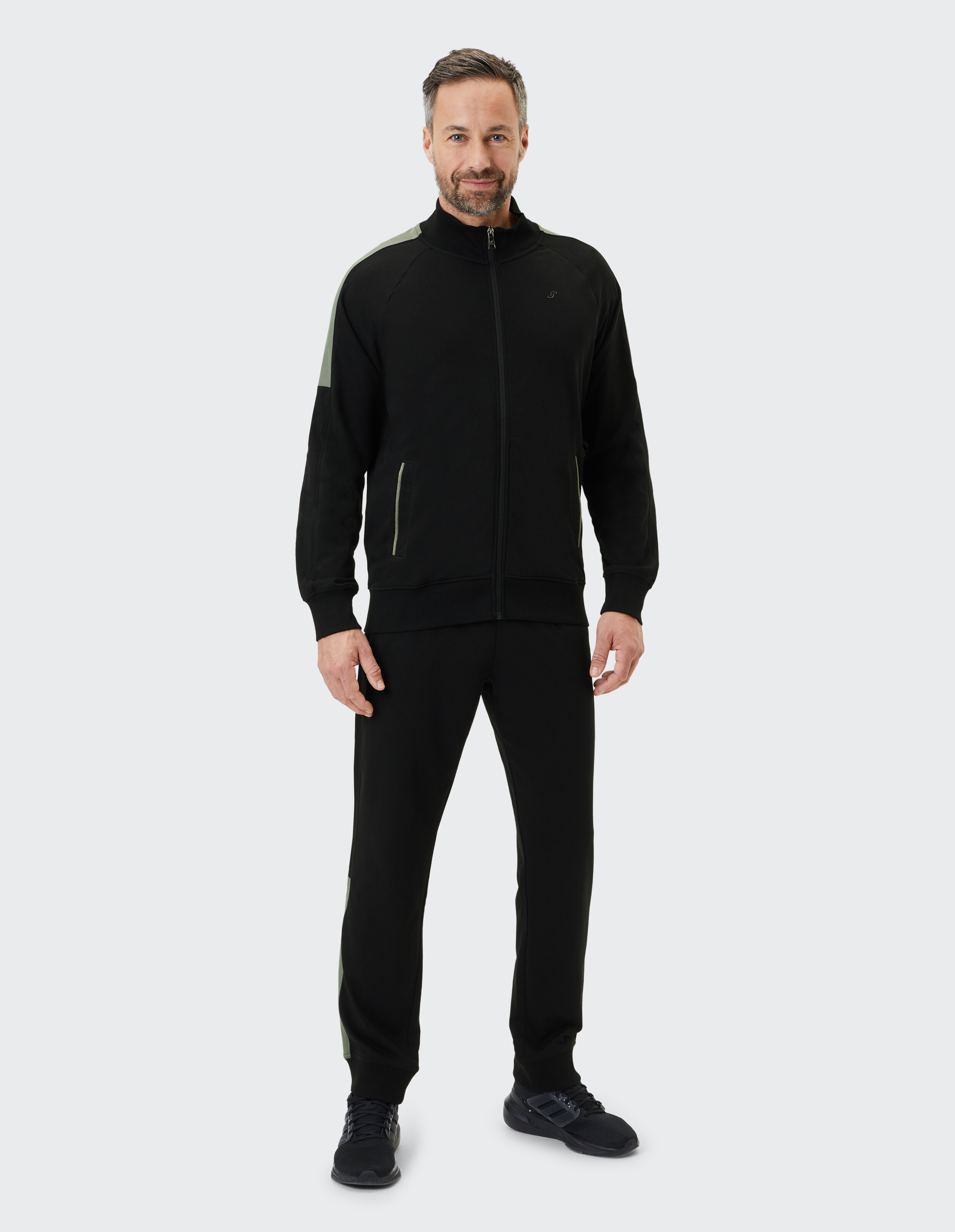 Schwarzer Trainingsanzug mit Reißverschlussjacke, grauen Details an Schultern und Hosenbeinen, sowie Seitentaschen.