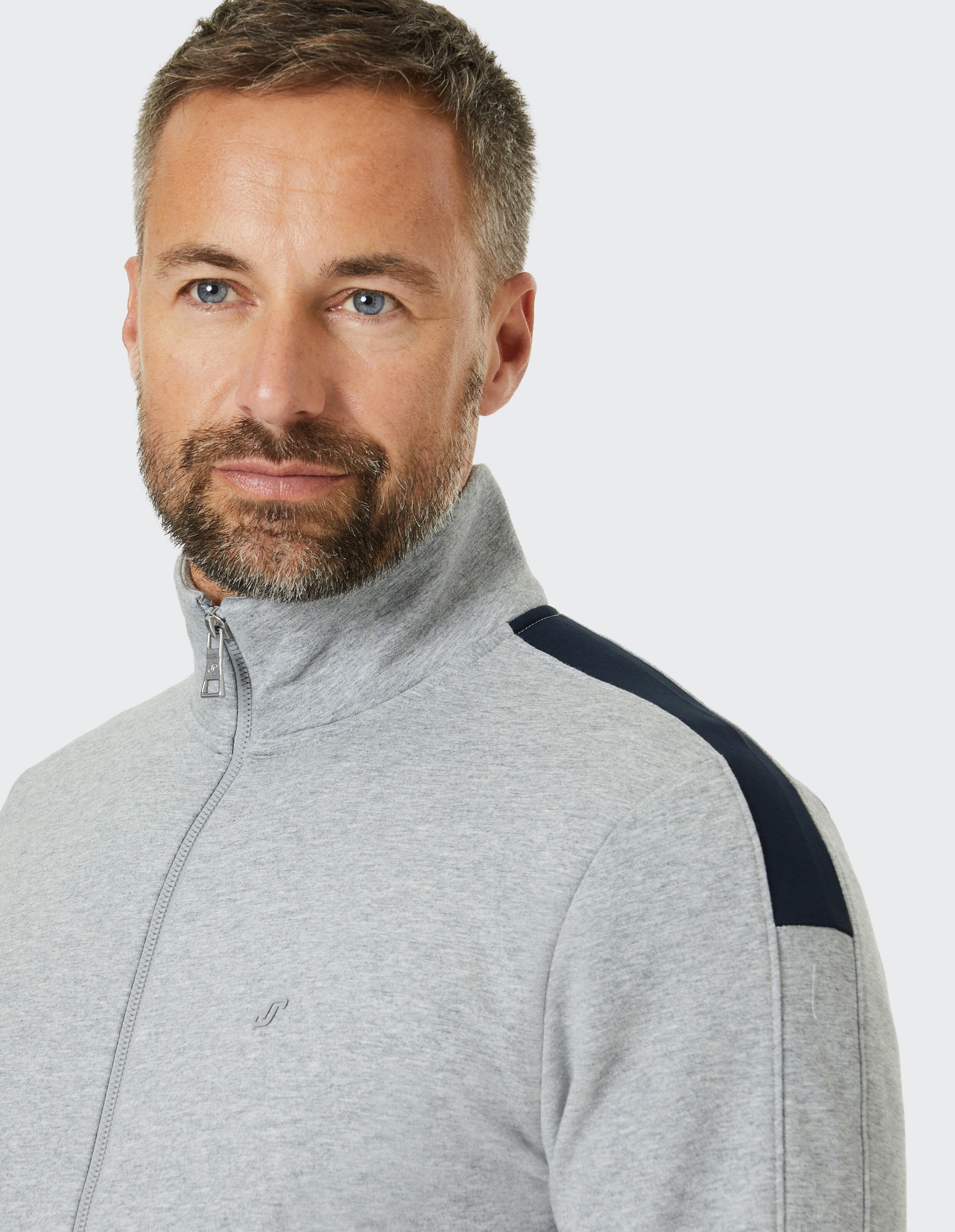Ein lächelnder Mann mit Bart in einer grau-blauen Jacke mit Reißverschluss und Stehkragen vor weißem Hintergrund.