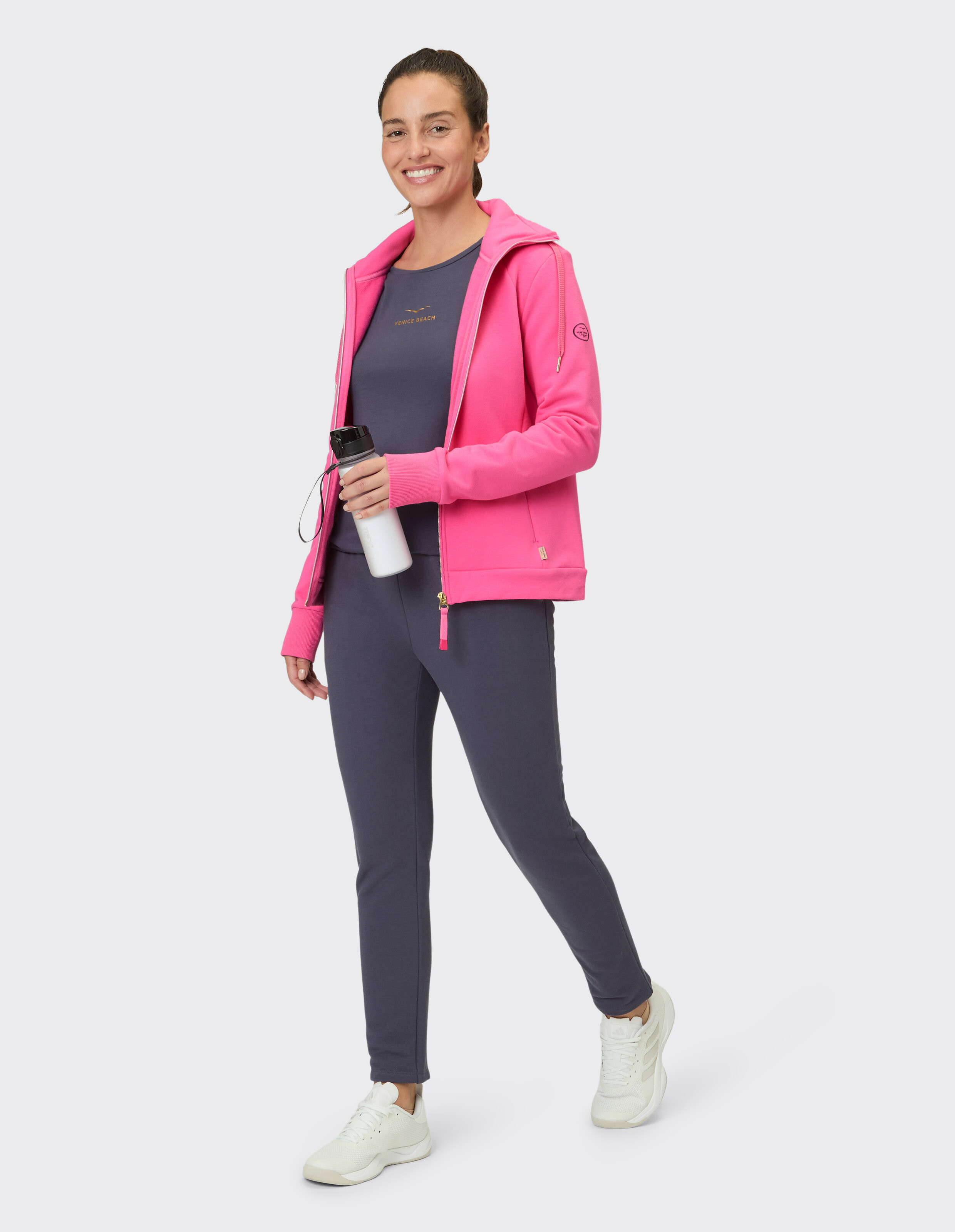 Frau trägt einen leuchtend rosa Kapuzenpullover mit Reißverschluss und ein dunkelgraues, lässiges Lounge-Set.