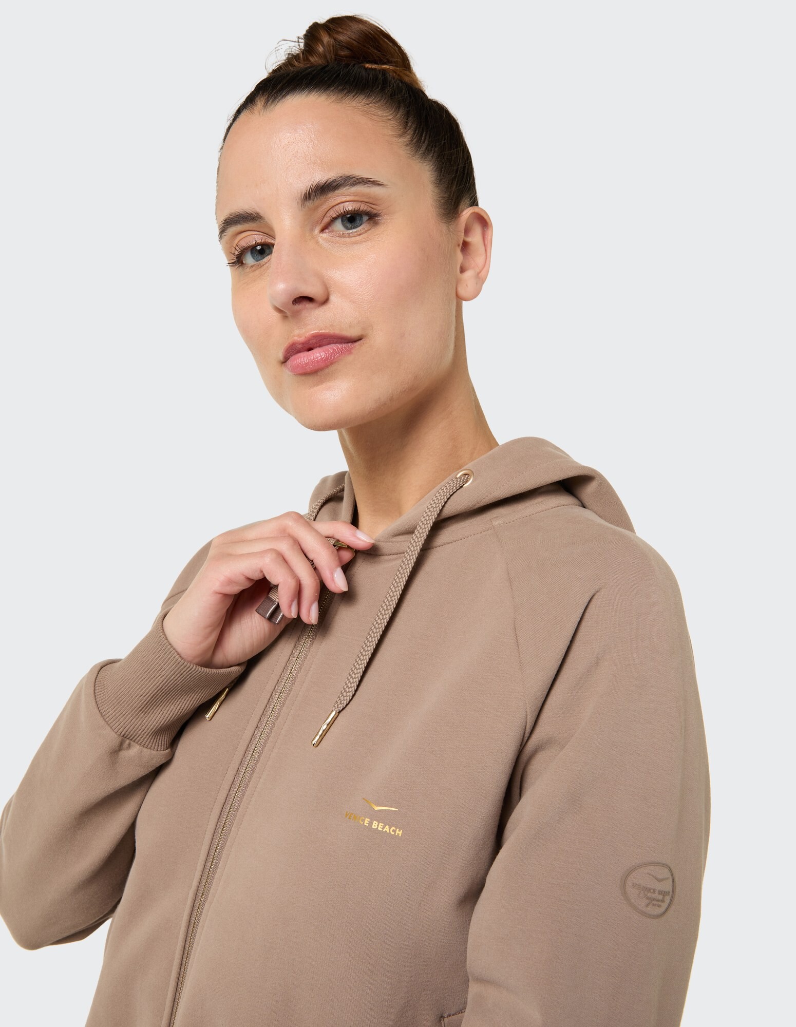 Nahaufnahme einer Frau in einem beigen Kapuzenpullover mit Reißverschluss und goldenem Logo, die einen lässigen Stil unterstreicht.