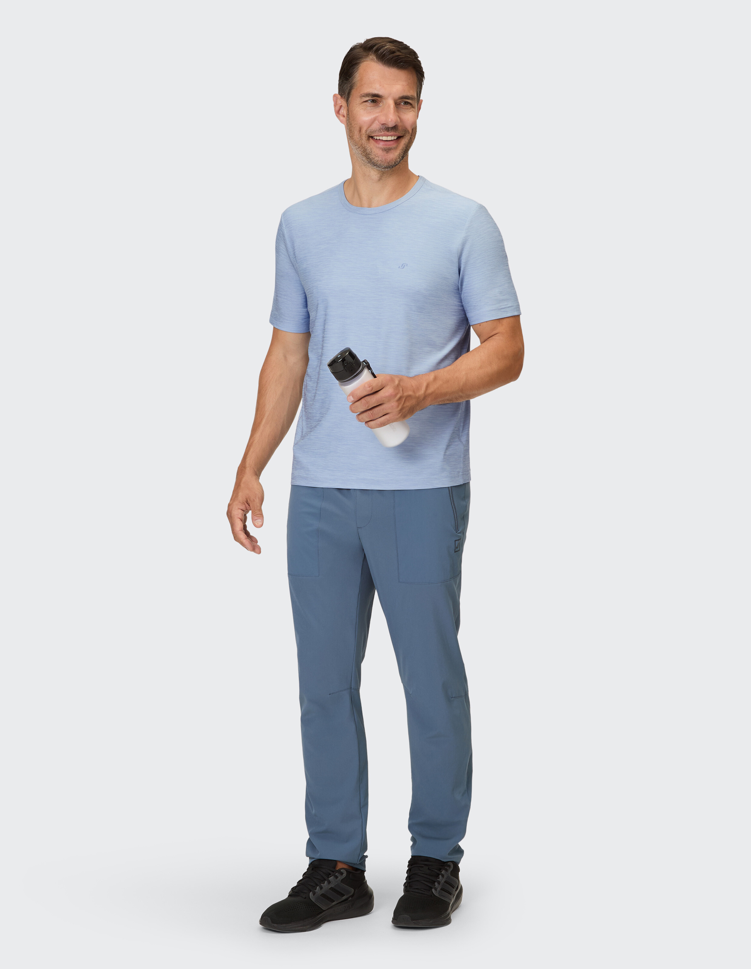 Ein Mann im hellblauen kurzärmeligen Shirt und blauen Cargohosen mit Seitentaschen hält eine weiße Trinkflasche.
