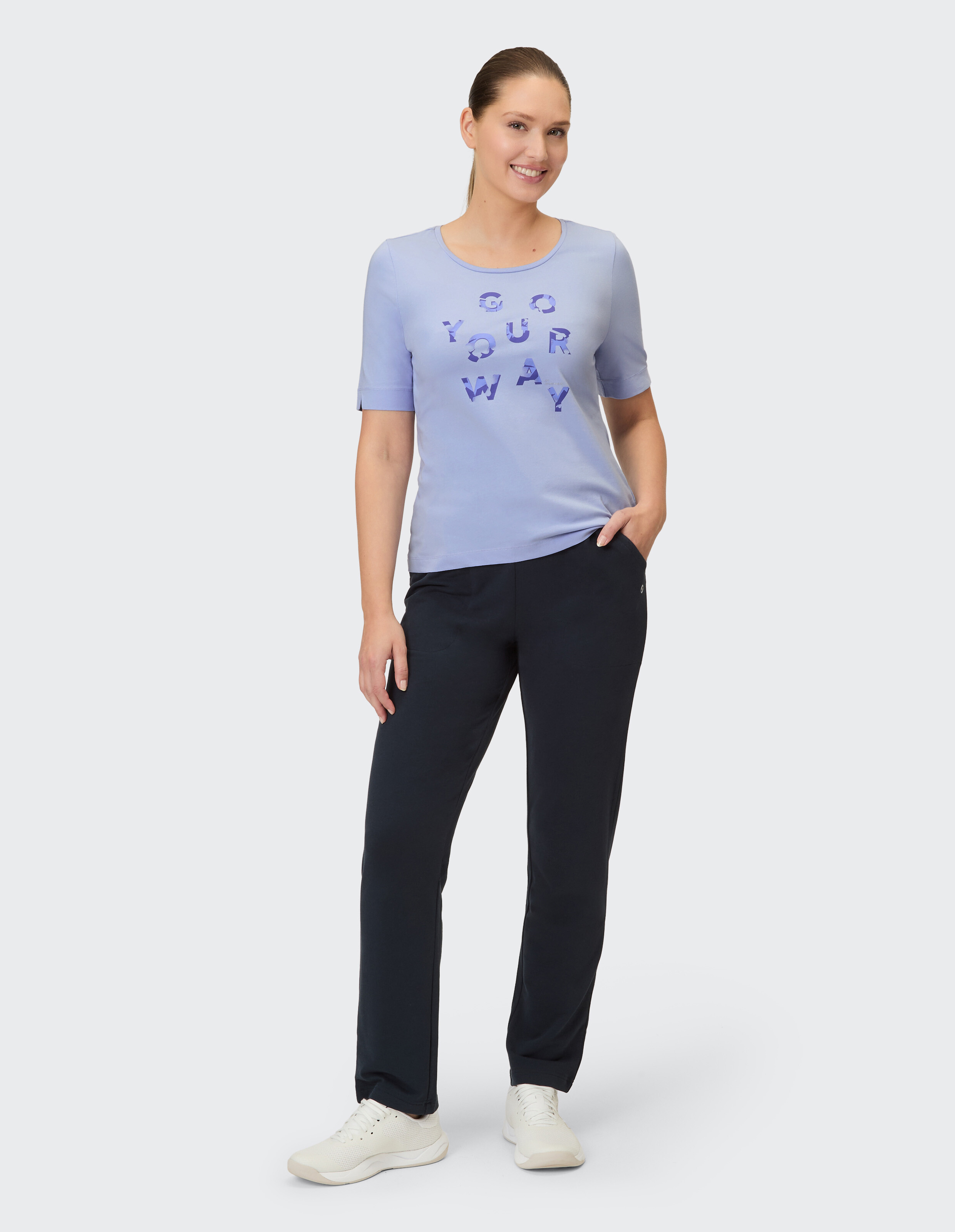 Eine Frau trägt ein hellblaues T-Shirt mit Schriftzug und dunkle Hose.