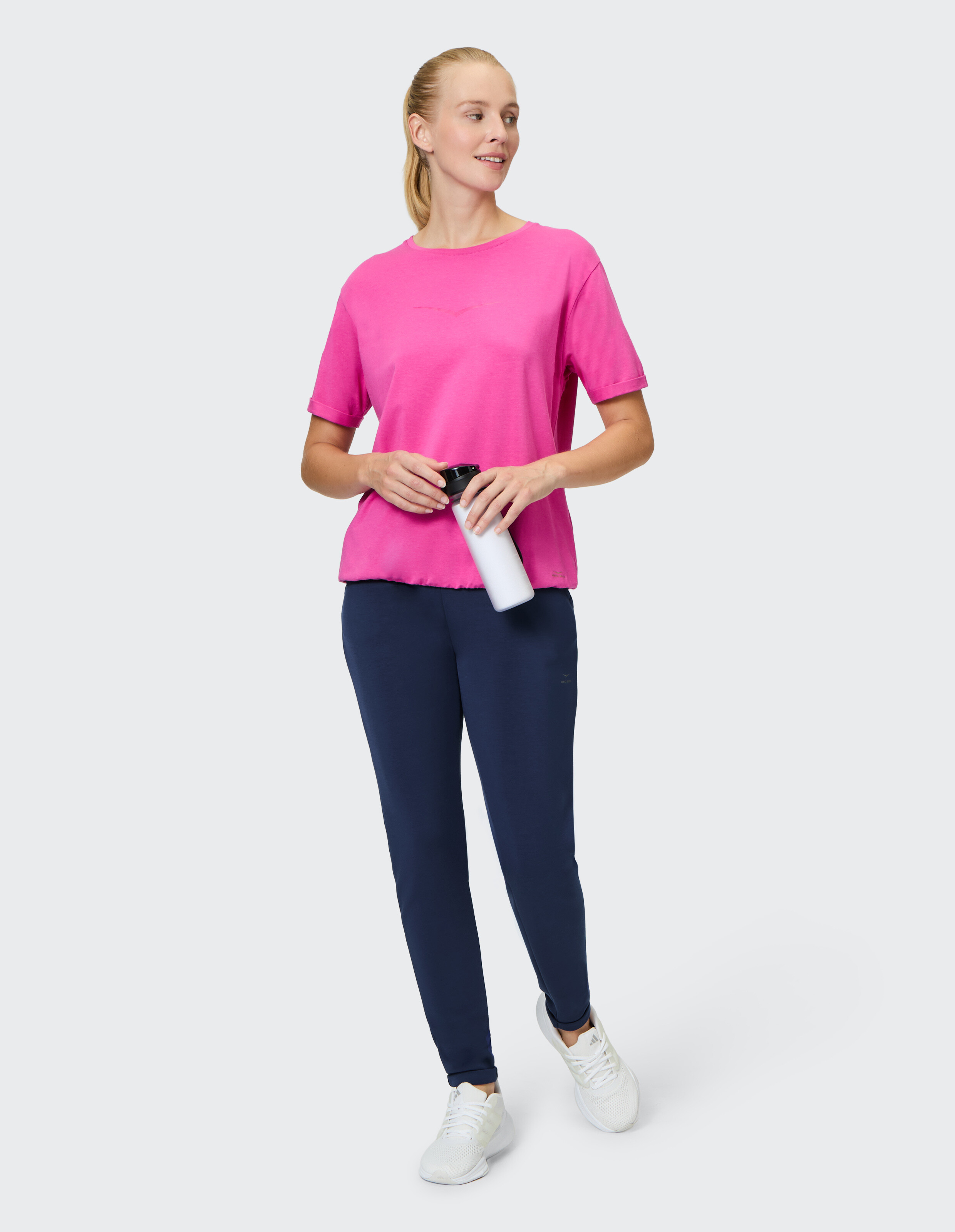 Frau in einem rosa T-Shirt und einer dunkelblauen Hose, eine Wasserflasche in der Hand, lässiger Stil.