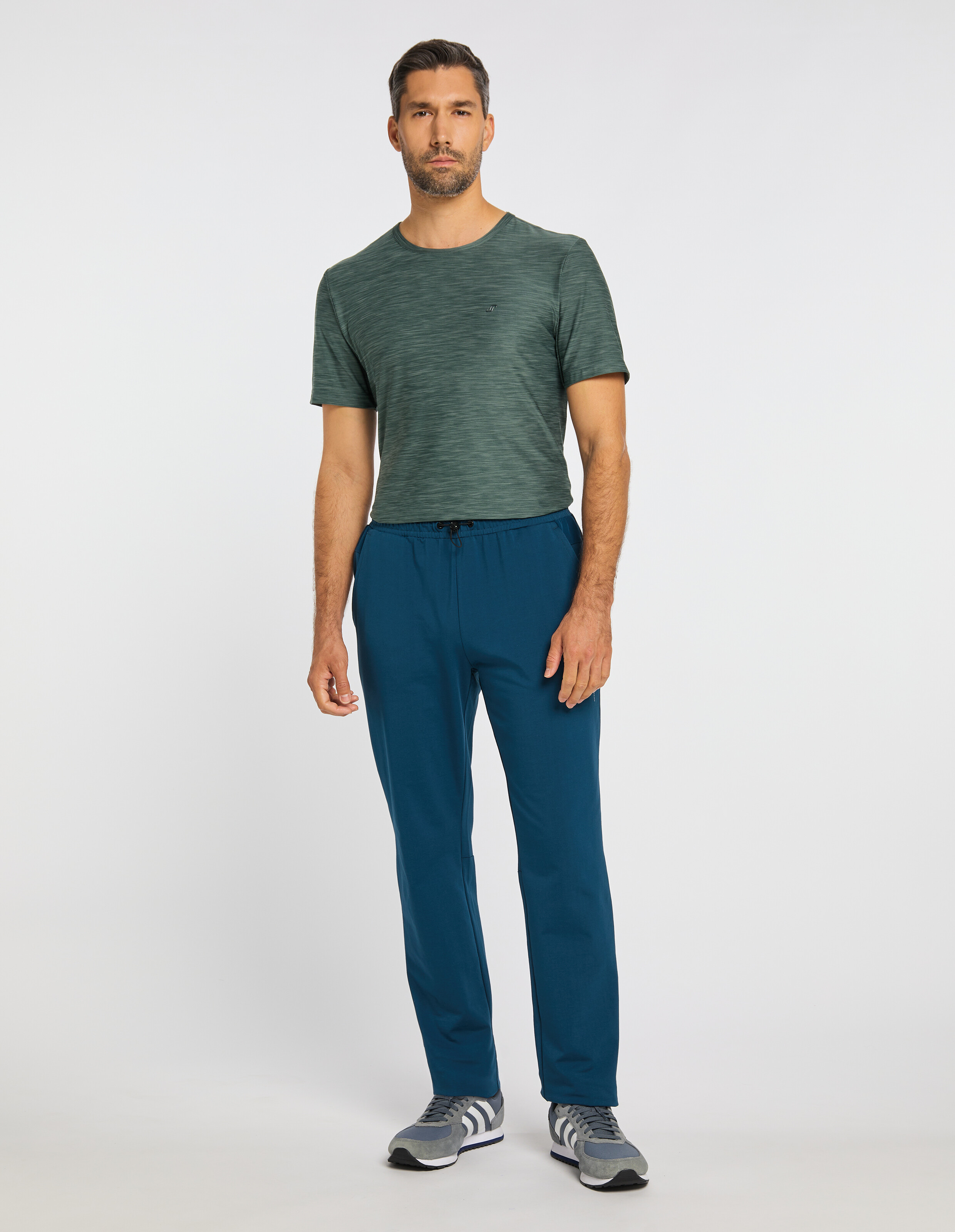 Ein Mann mit Bart trägt ein grünes T-Shirt, blaue Jogginghose und graue Sneakers vor einem neutralen, weißen Hintergrund.