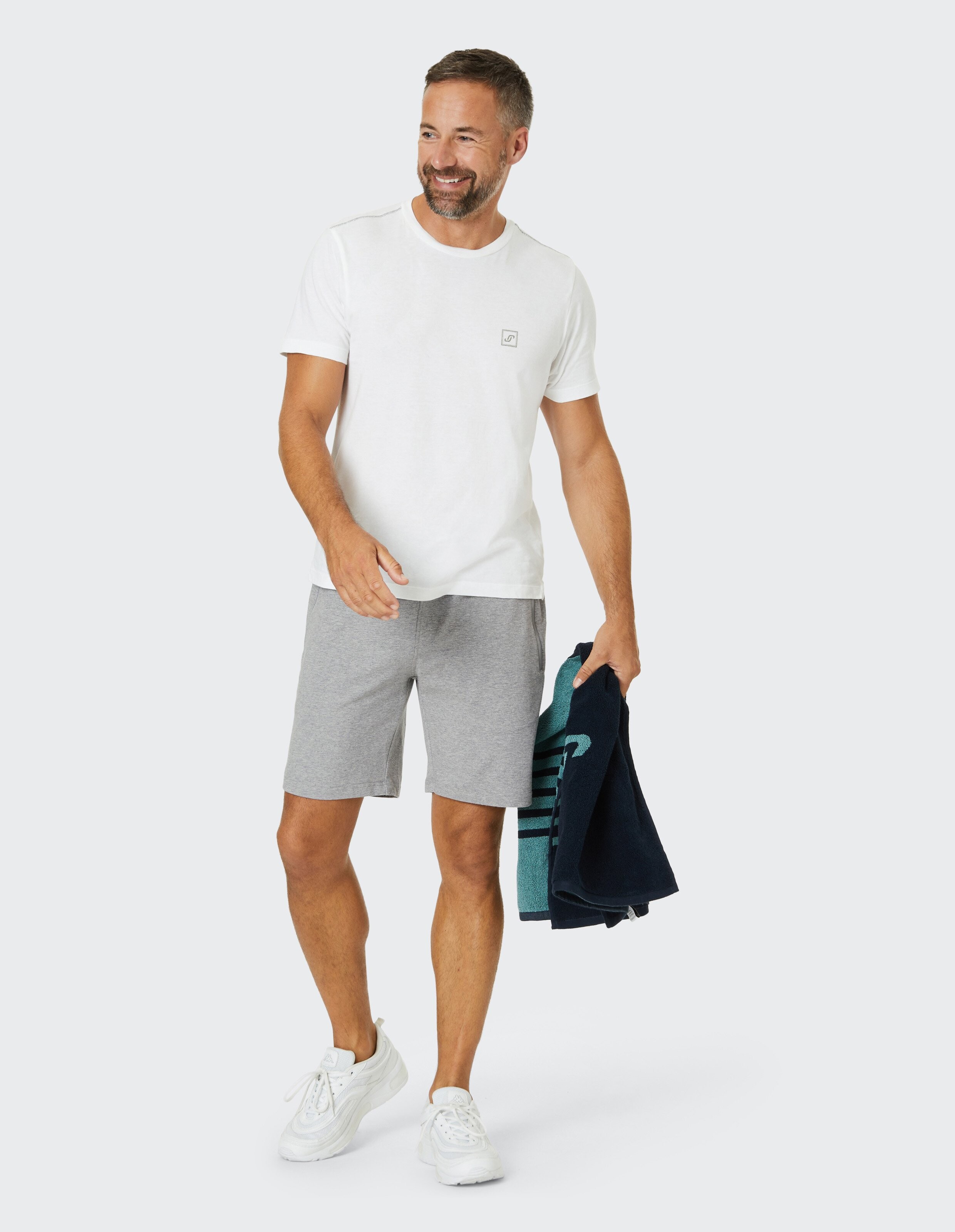 Ein lächelnder Mann trägt ein weißes T-Shirt und graue Shorts, hält eine gefaltete Jacke, steht casual in weißen Sneakers.