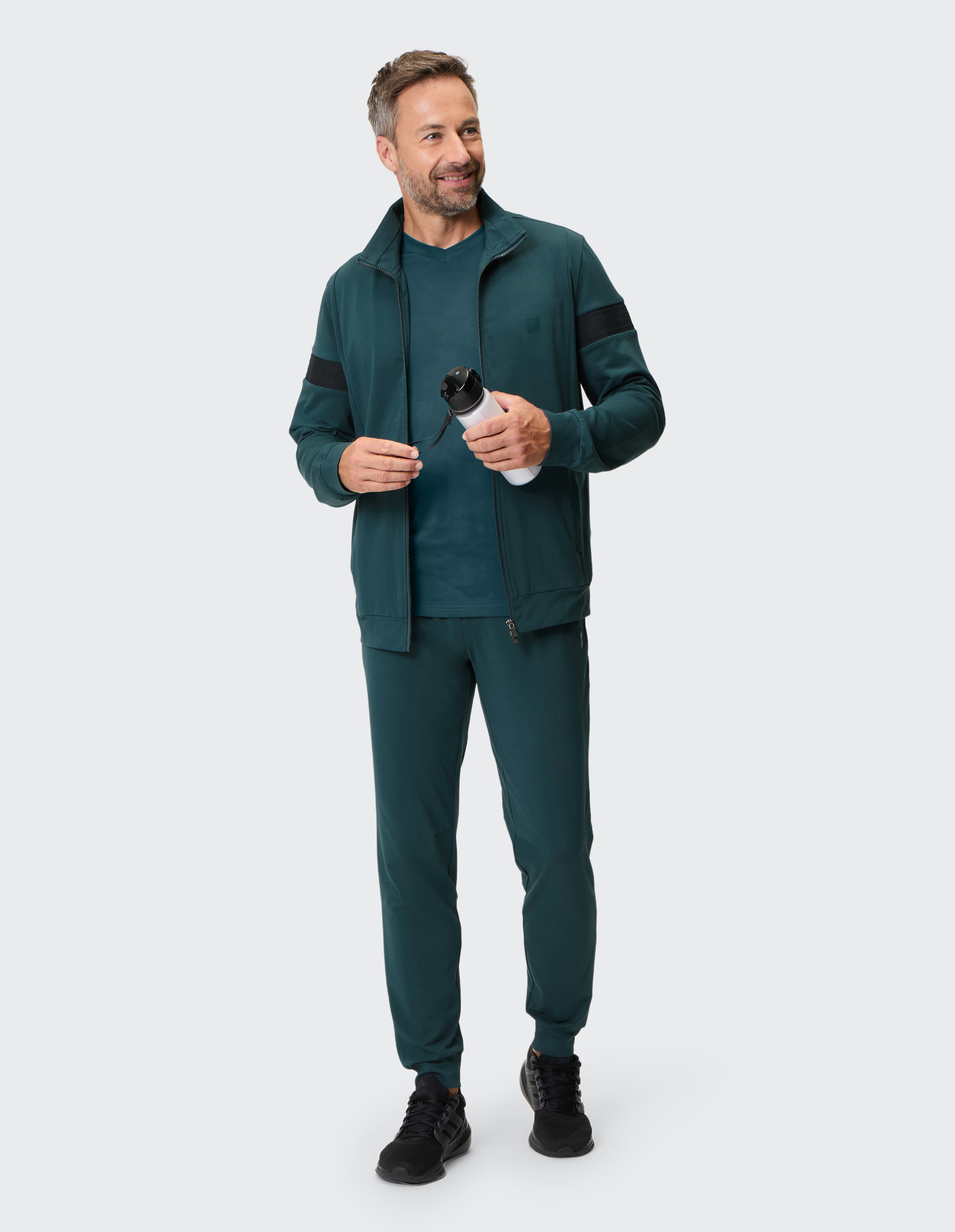 Mann trägt einen dunkelgrünen Freizeit-Trainingsanzug mit einer Jacke mit Reißverschluss und passender Jogginghose.