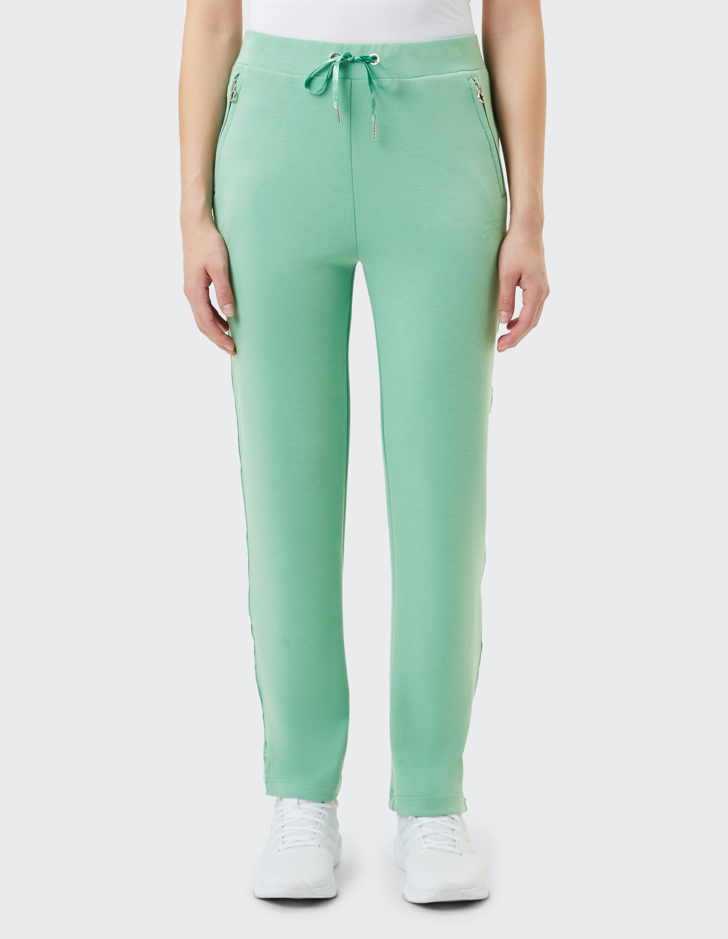 Eine Person trägt eine grüne Jogginghose mit Kordelzug und Reißverschlusstaschen, dazu weiße Sportschuhe und ein weißes Oberteil.