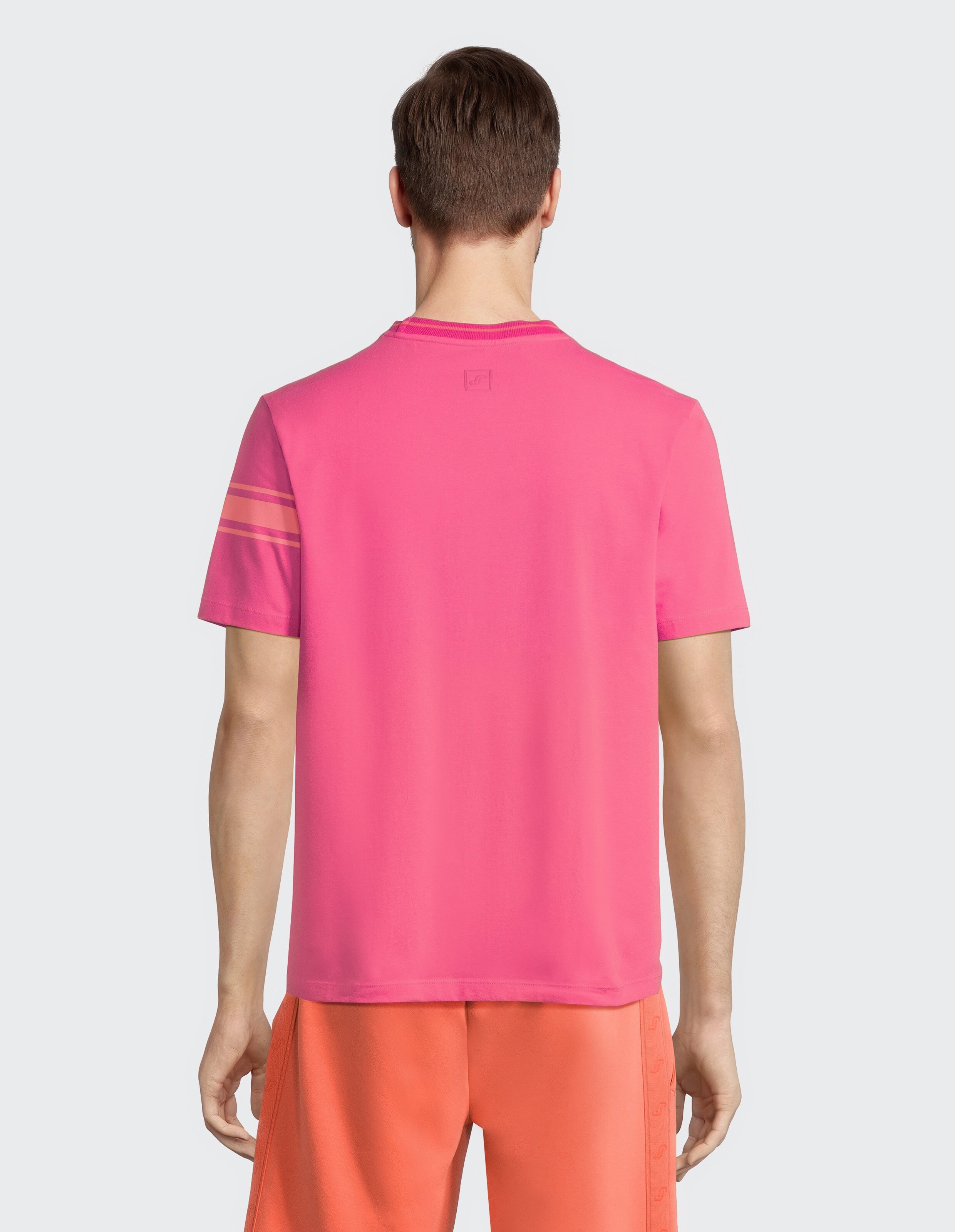 Ein Mann steht mit dem Rücken zur Kamera, trägt ein pinkes T-Shirt und orangefarbene Shorts vor neutralem Hintergrund.