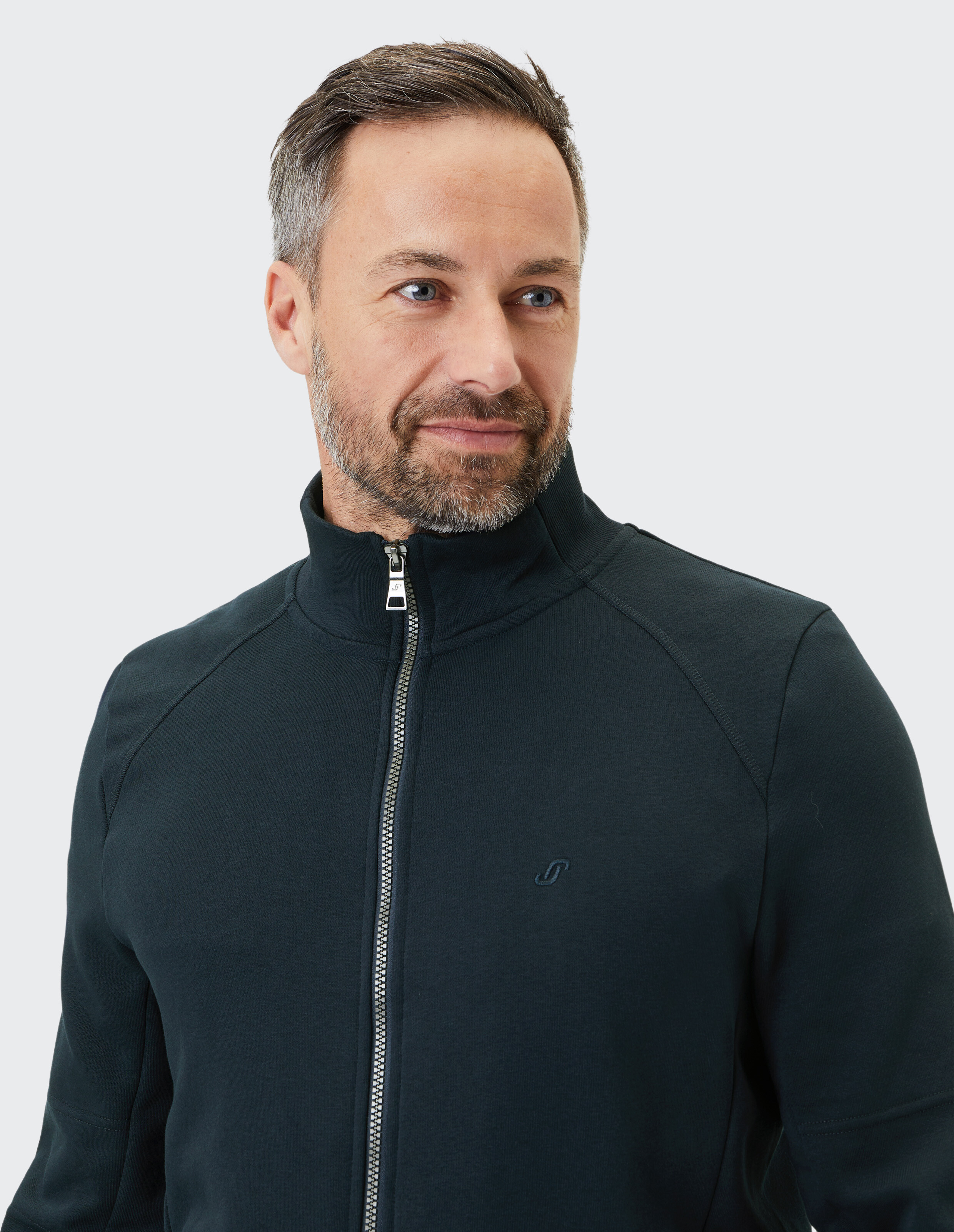 Ein Mann mit kurzem Haar und Bart trägt eine schwarze, sportliche Jacke mit Reißverschluss vor einem grauen Hintergrund.