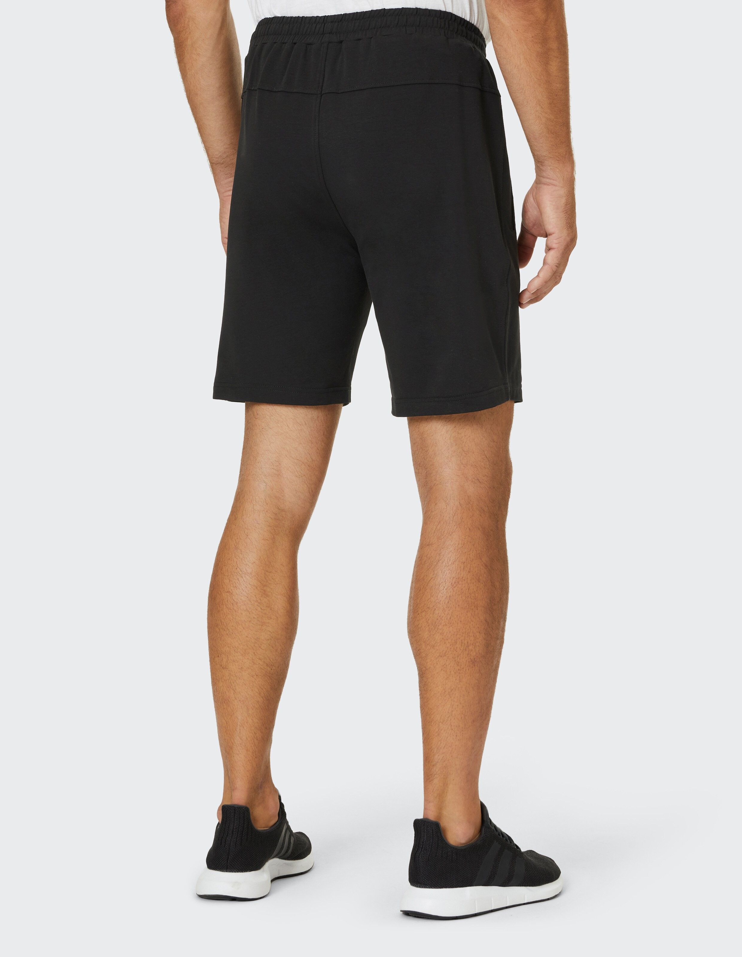 Ein Mann trägt schwarze Shorts und schwarze Sneaker auf weißem Hintergrund, von hinten fotografiert; trägt ein weißes Oberteil.