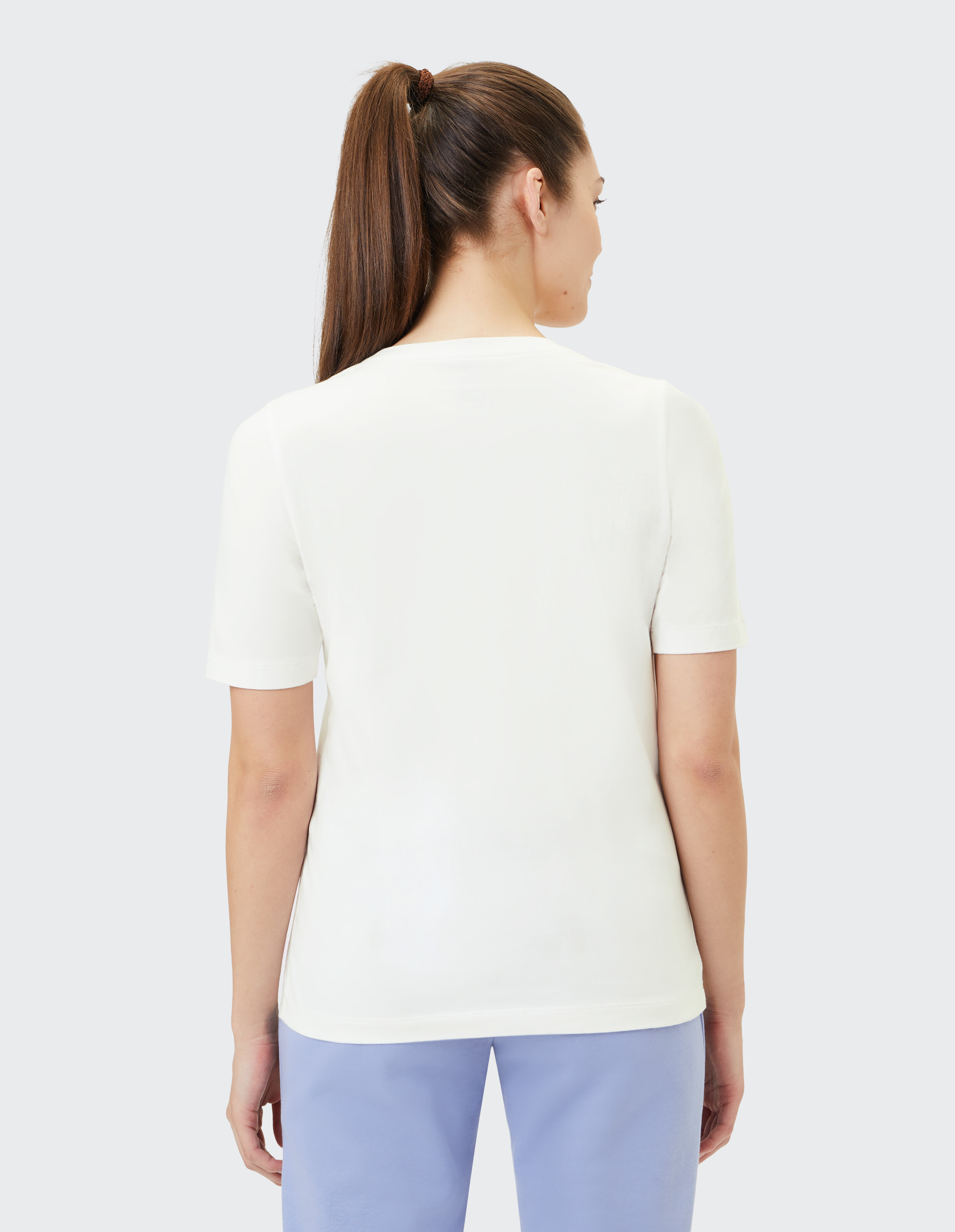 Frau mit langen Haaren im Pferdeschwanz trägt ein weißes T-Shirt und hellblaue Hose, steht mit dem Rücken zur Kamera vor grauem Hintergrund.