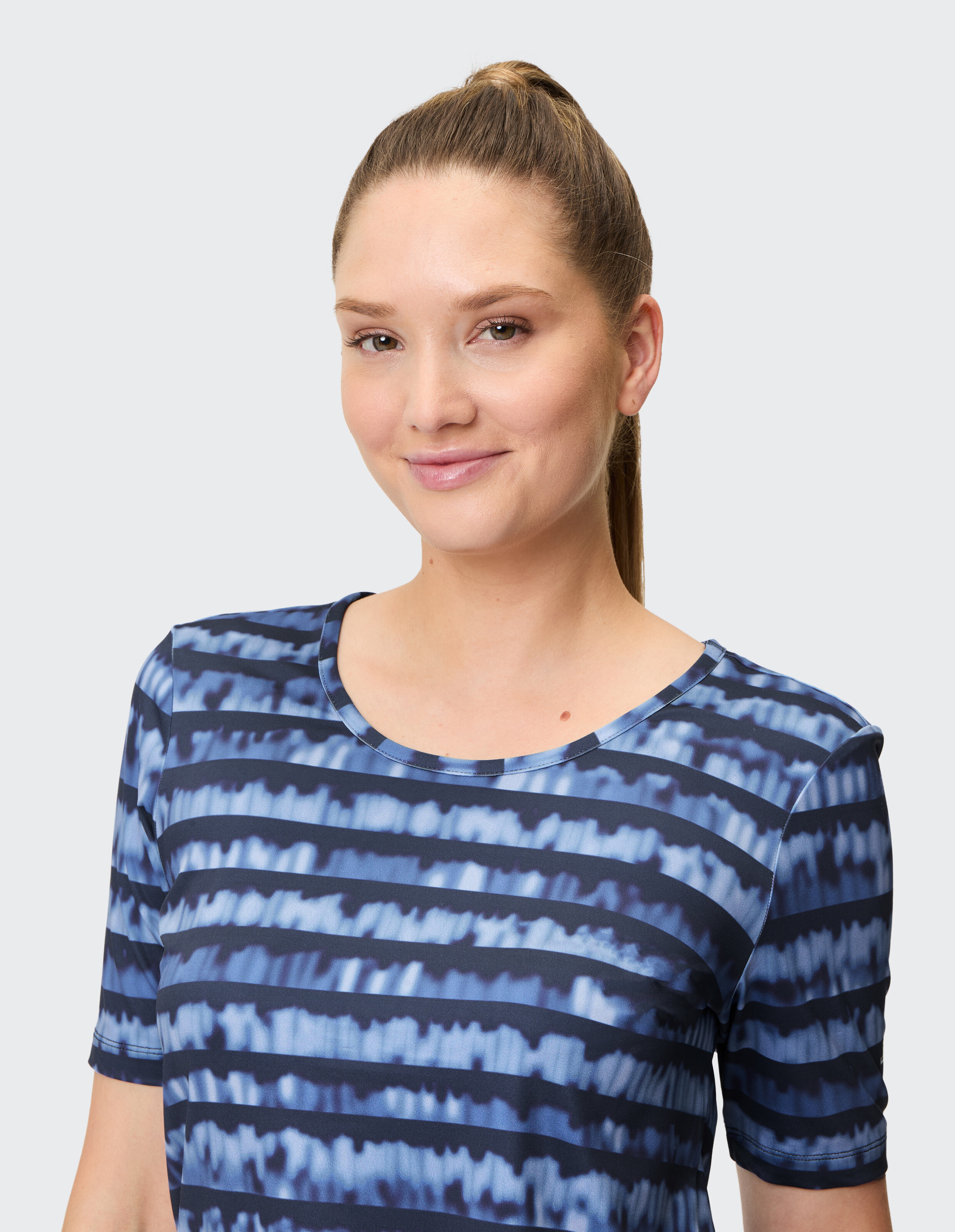 Frau mit zurückgebundenem Haar trägt ein blau-gestreiftes Shirt und lächelt dezent vor einem grauen Hintergrund.