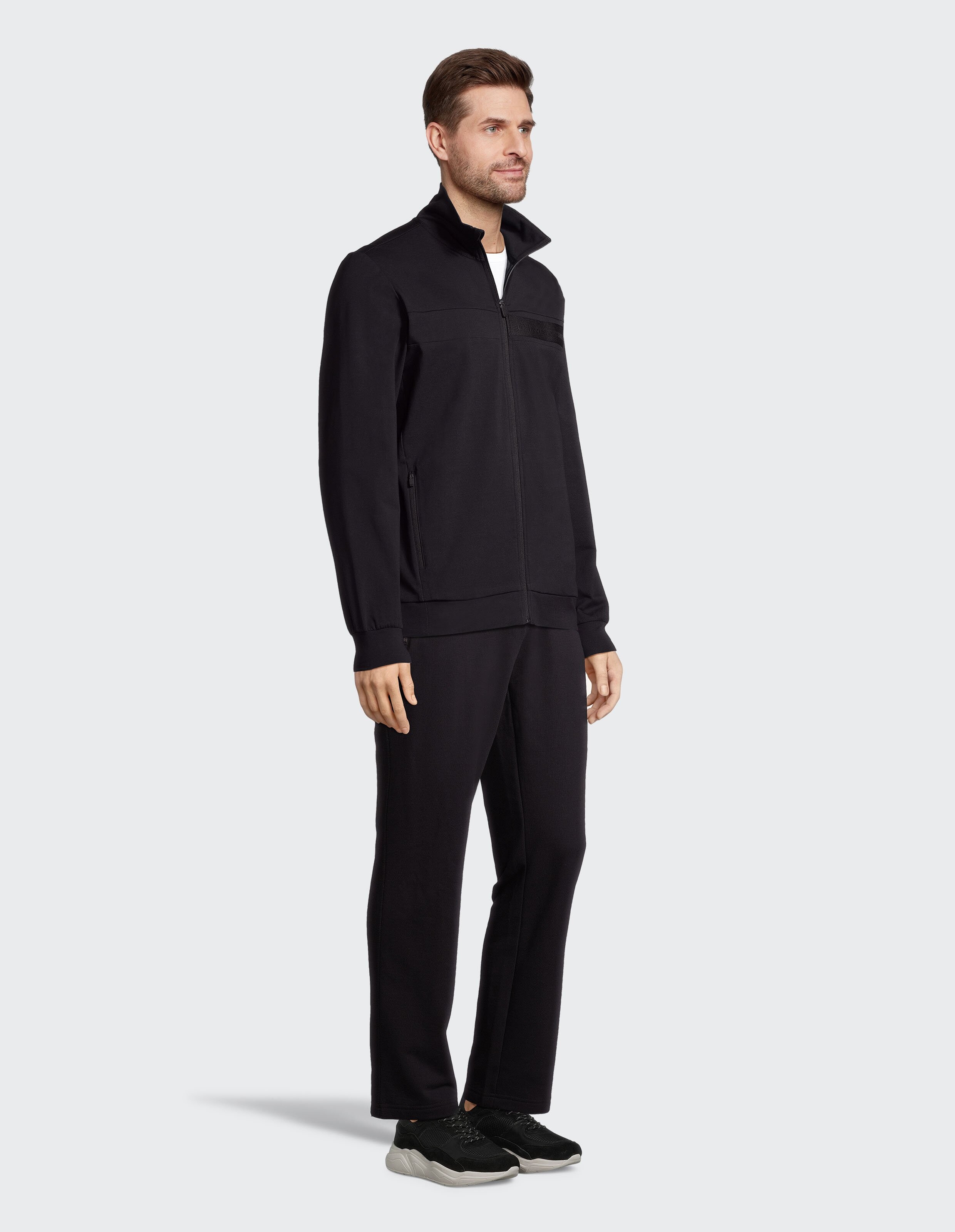 Ein Mann in schwarzer Jogginghose und schwarzer Jacke mit Reißverschluss steht seitlich, trägt schwarze Sneakers auf grauem Hintergrund.