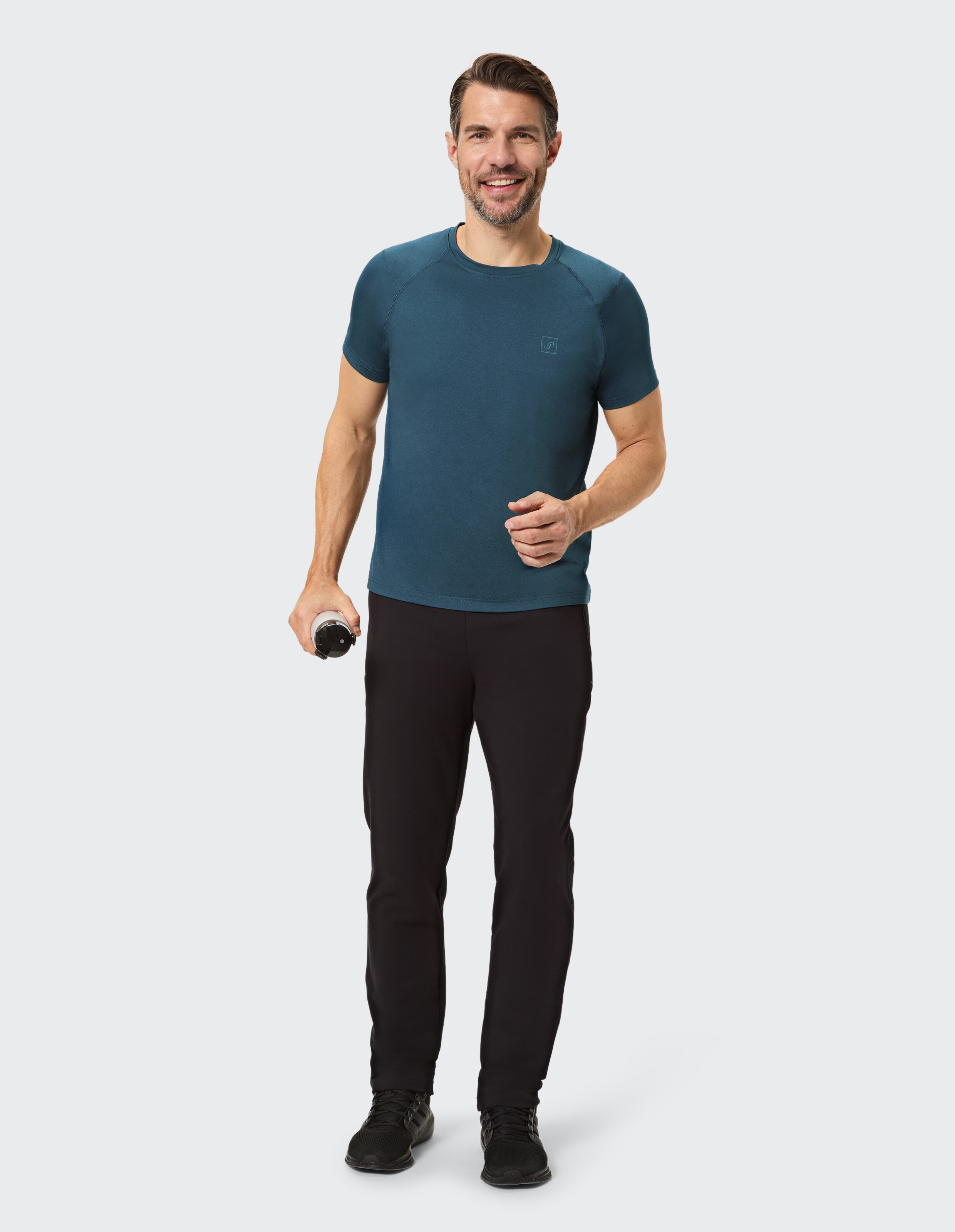 Mann in blaugrünem Kurzarmhemd und schwarzer Hose, mit einer Wasserflasche in der Hand.