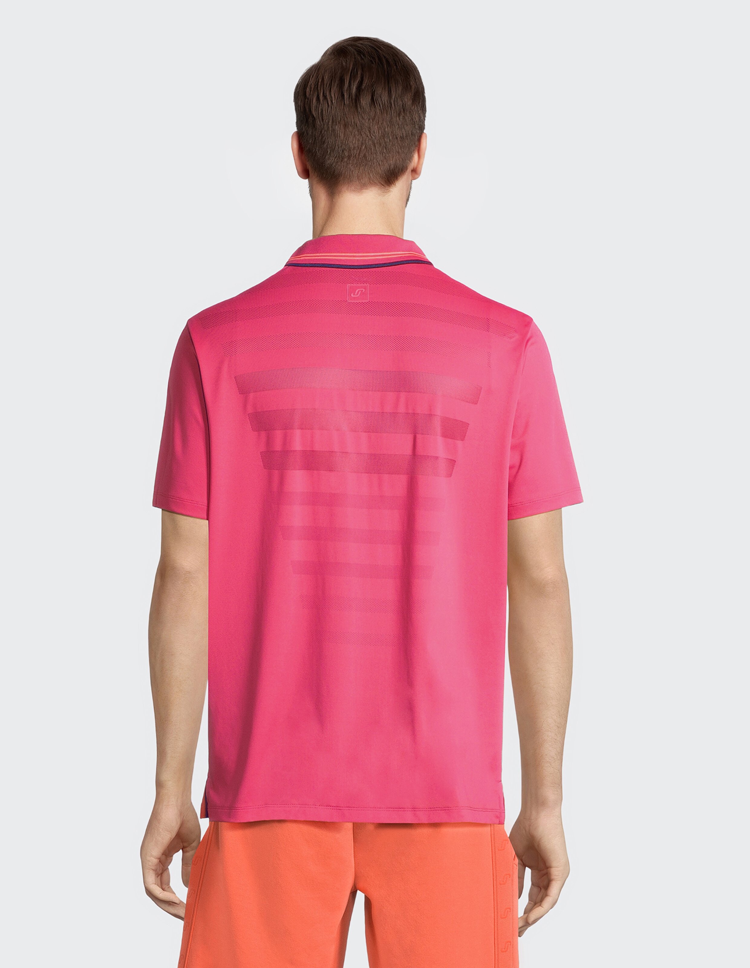 Eine Person steht mit dem Rücken zur Kamera, trägt ein pinkfarbenes Polohemd mit einem gestreiften Muster und orange Shorts.