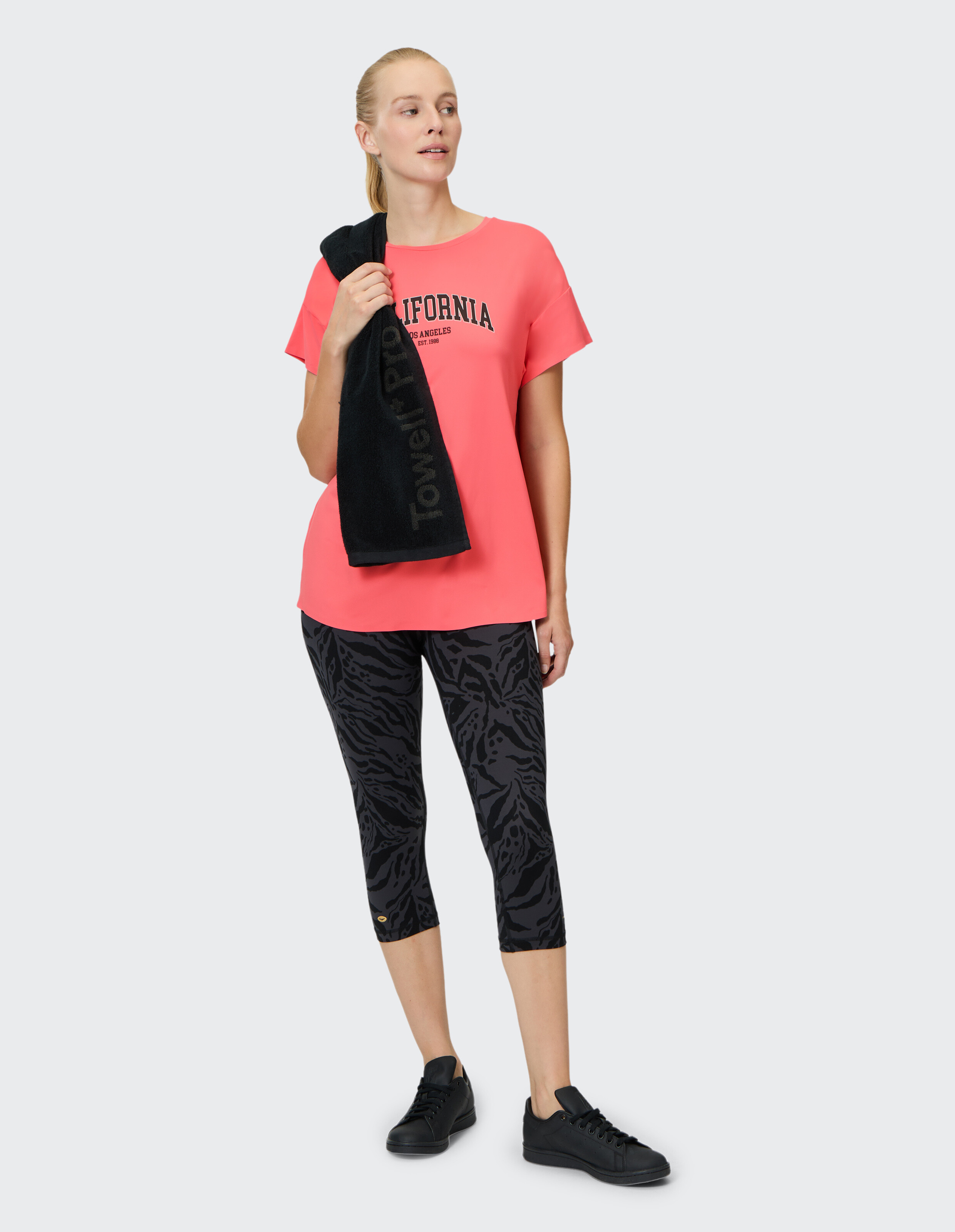Frau trägt ein korallenrotes Venice Beach Kurzarmshirt Edana und gemusterte Capri-Leggings.