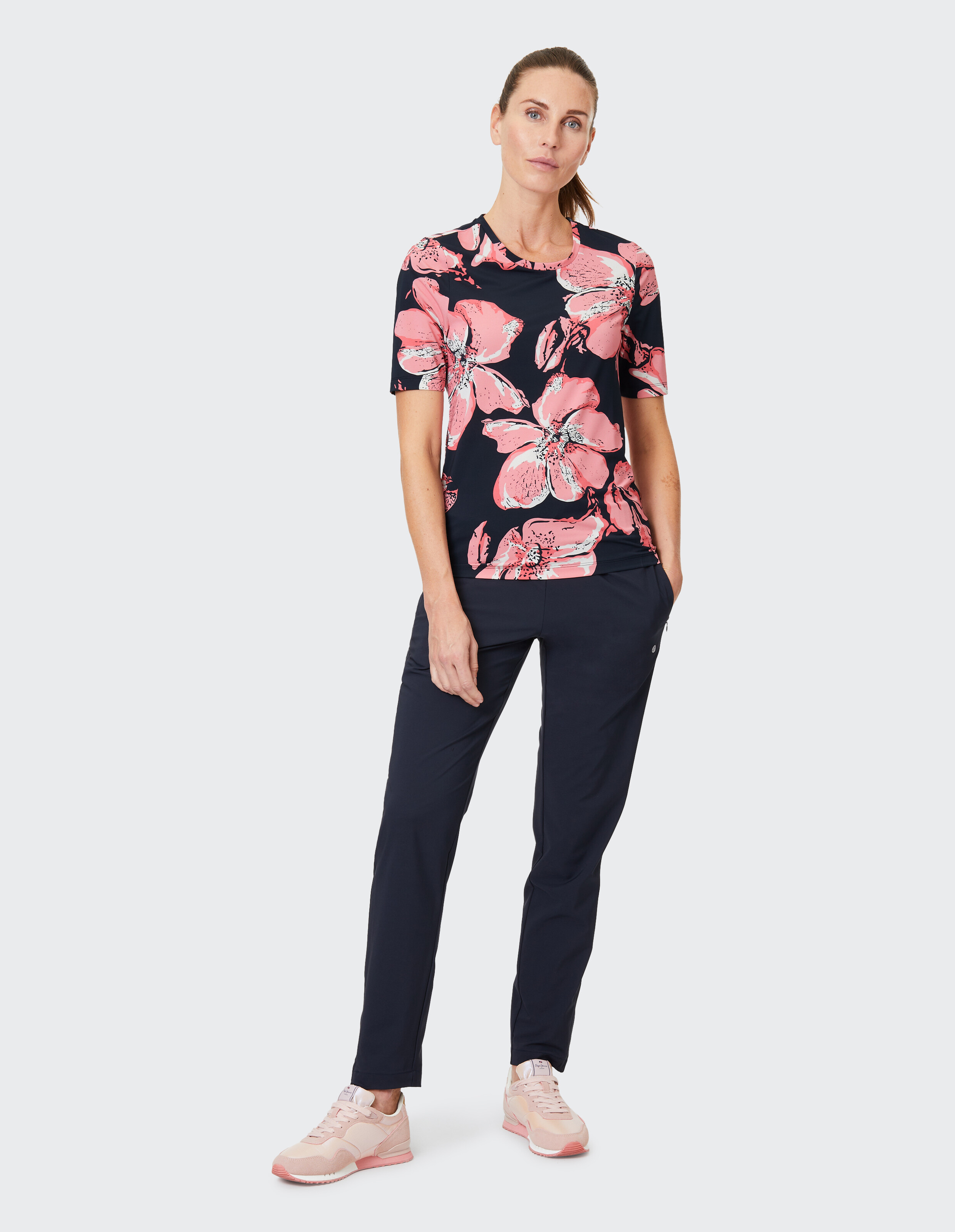 Frau trägt ein dunkles T-Shirt mit großen rosa Blumenmustern, dunkle Hose und rosa Sportschuhe vor neutralem Hintergrund.