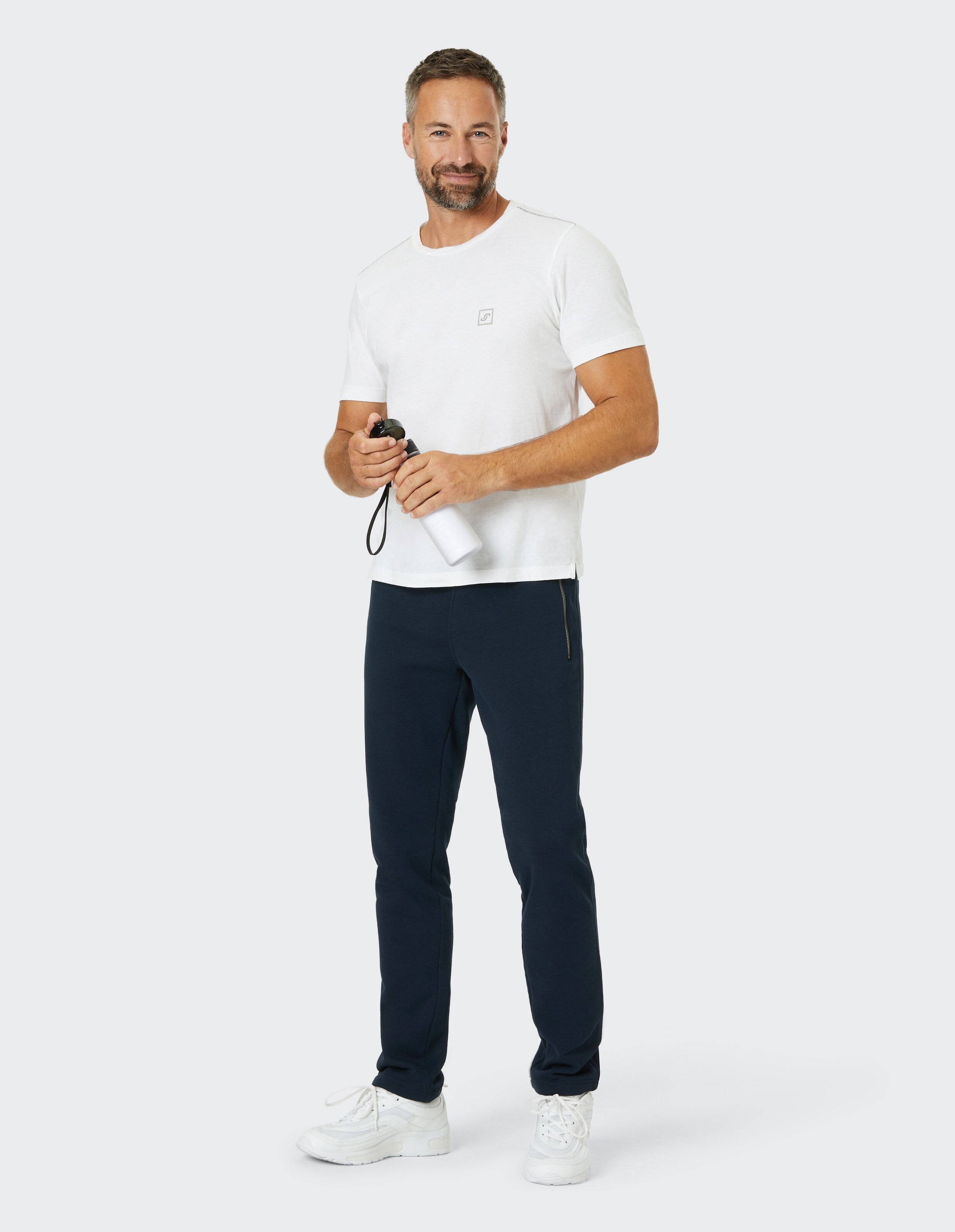 Ein Mann in weißem T-Shirt und dunkler Hose hält eine Wasserflasche und lächelt, während er locker in weißen Sneakers steht.