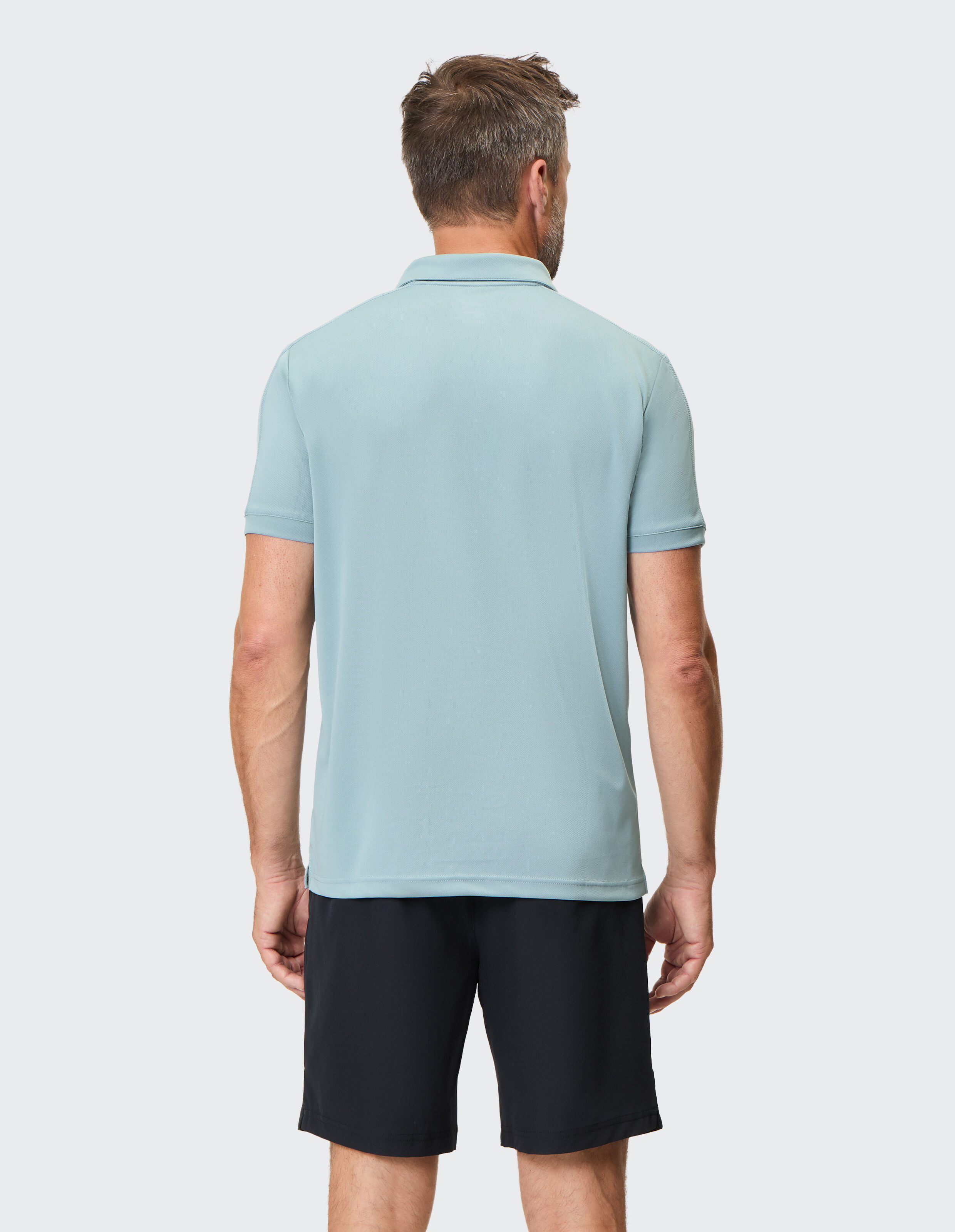 Rückansicht eines Mannes in hellblauem Poloshirt und schwarzen Shorts, stehend vor einem neutralen Hintergrund.