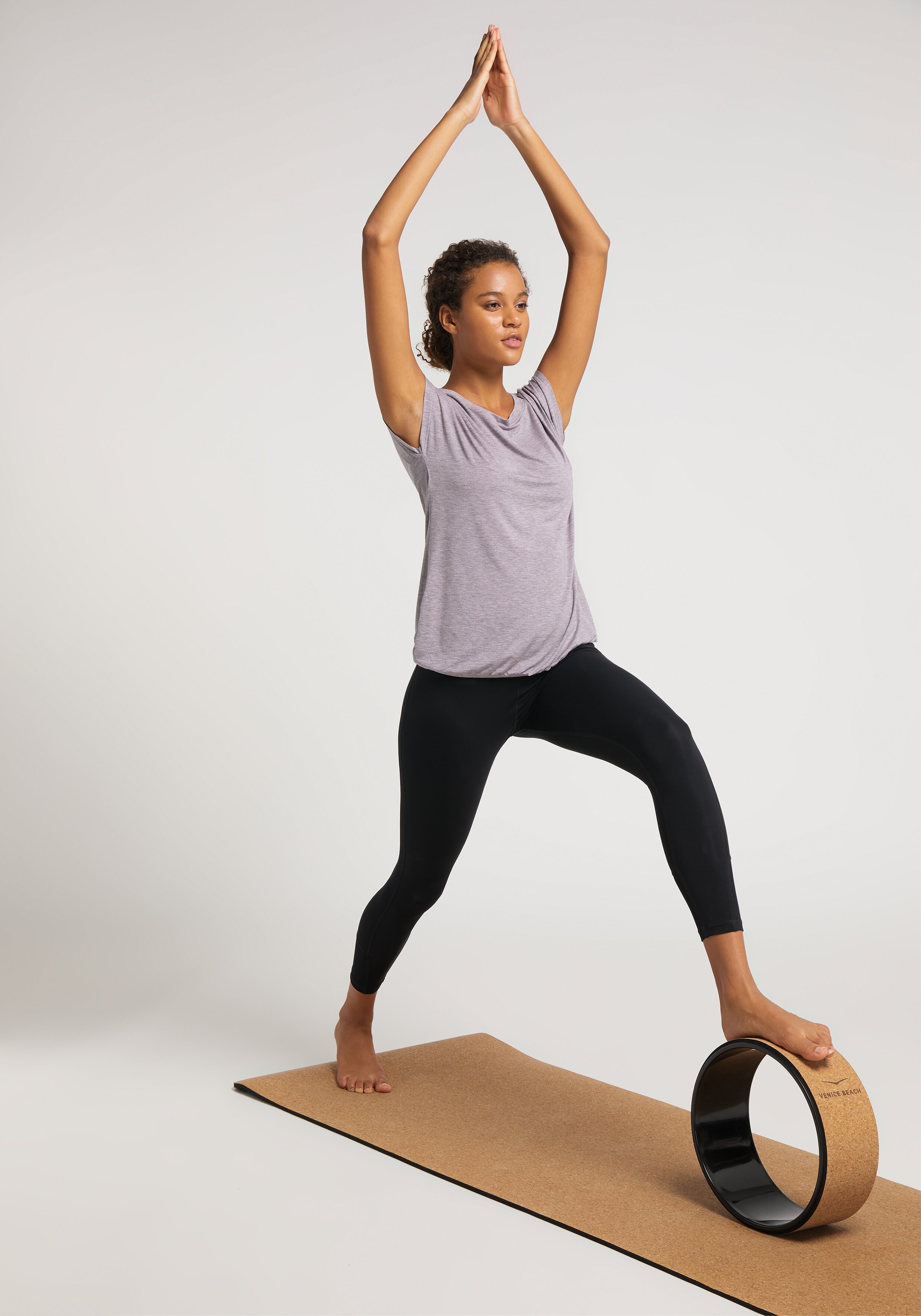 Frau in einem fliederfarbenen Top und schwarzen Leggings macht Yoga auf einer Korkmatte mit einem Rad.