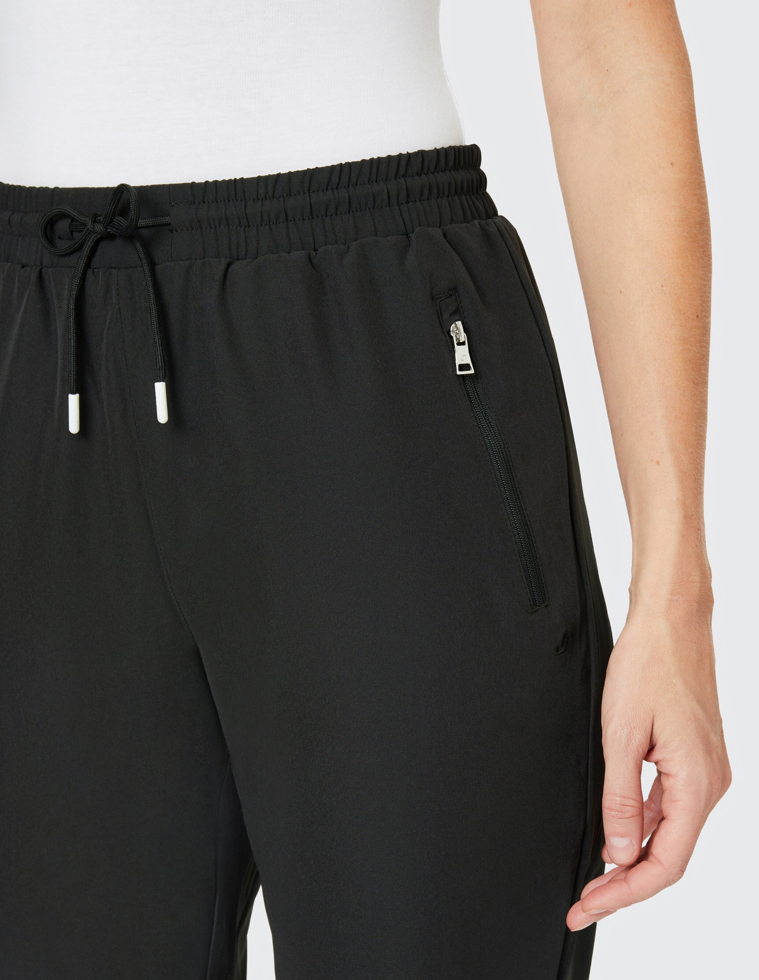 Eine Person trägt eine schwarze Jogginghose mit Kordelzug und Reißverschlusstasche, kombiniert mit einem weißen Oberteil.