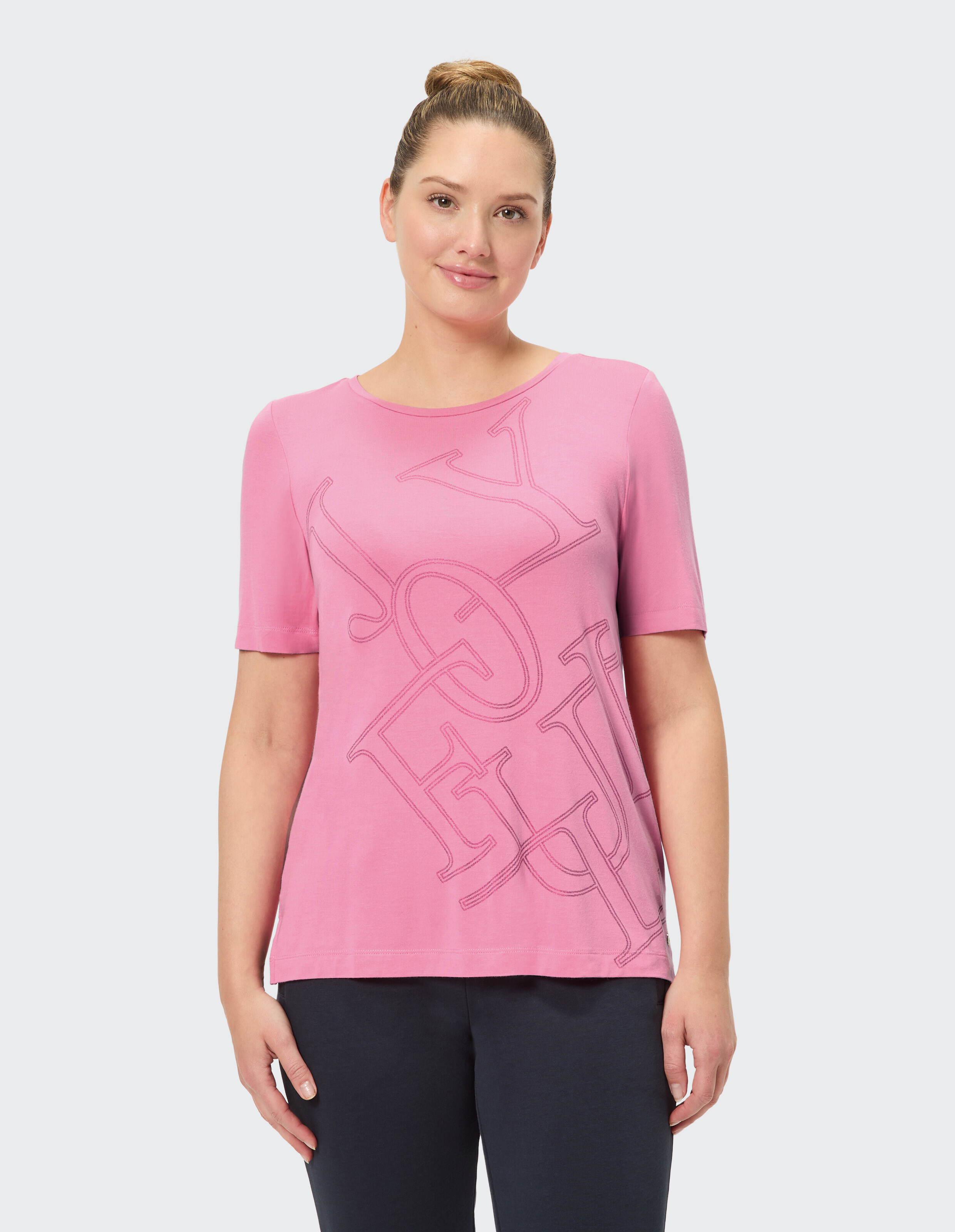 Eine lächelnde Person trägt ein pinkes T-Shirt mit großen, schrägen Buchstaben und einfachem Schnitt vor hellem Hintergrund.