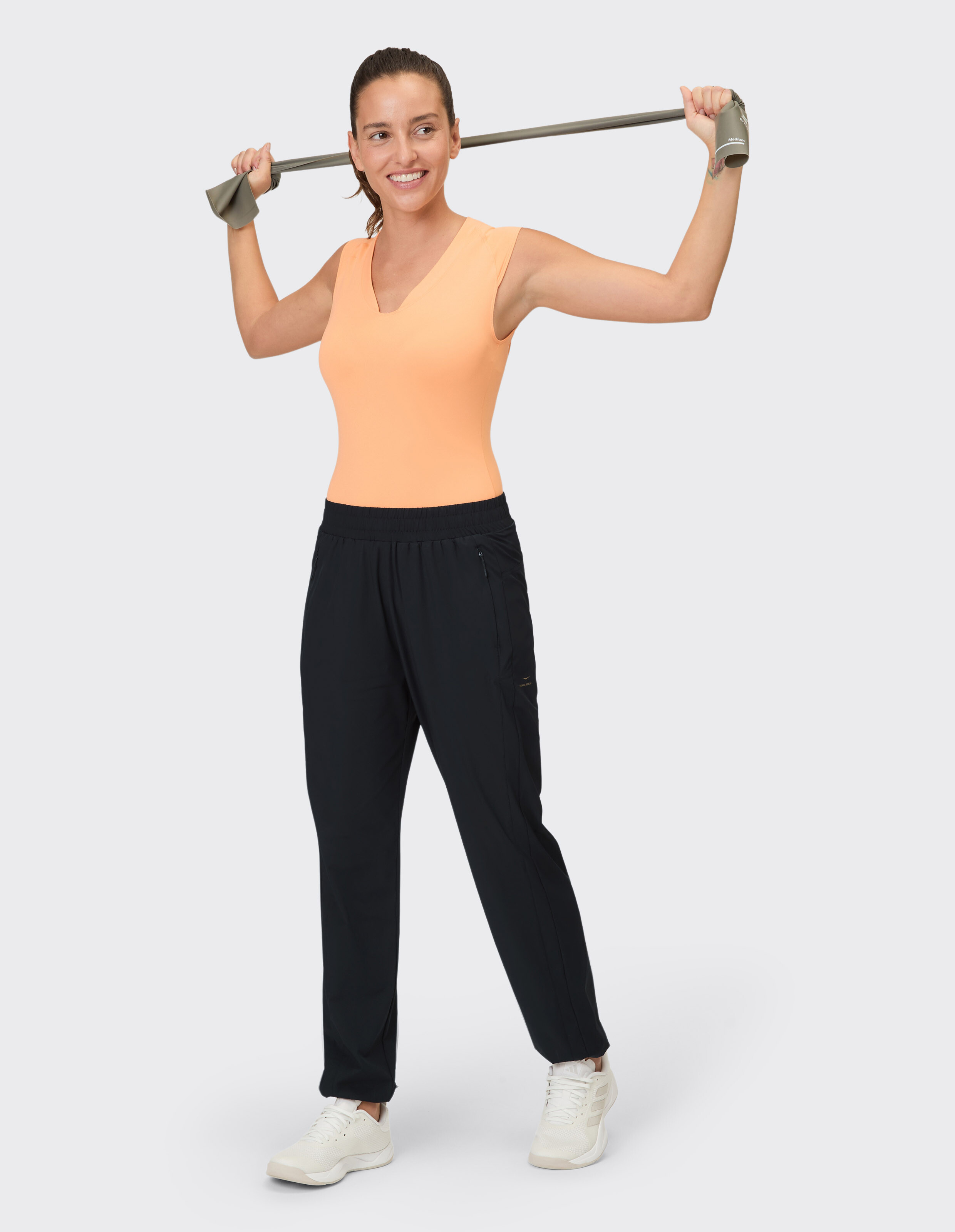 Frau in pfirsichfarbenem Tanktop und schwarzer Freizeithose trainiert mit einem Widerstandsband.
