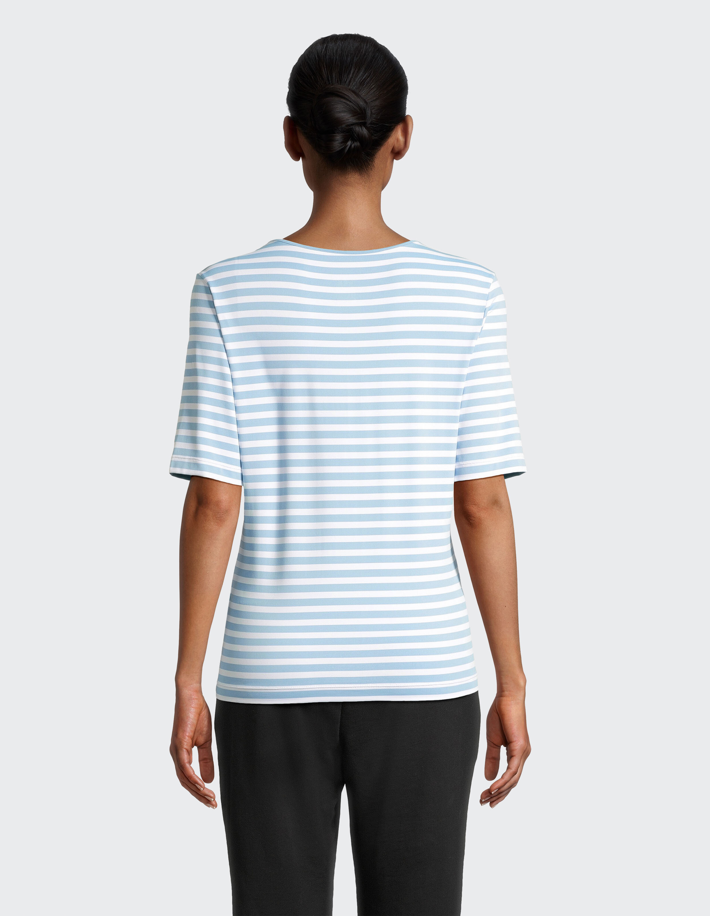 Person von hinten in einem hellblau-weiß gestreiften T-Shirt und schwarzen Hosen, die Haare zu einem Knoten gebunden.