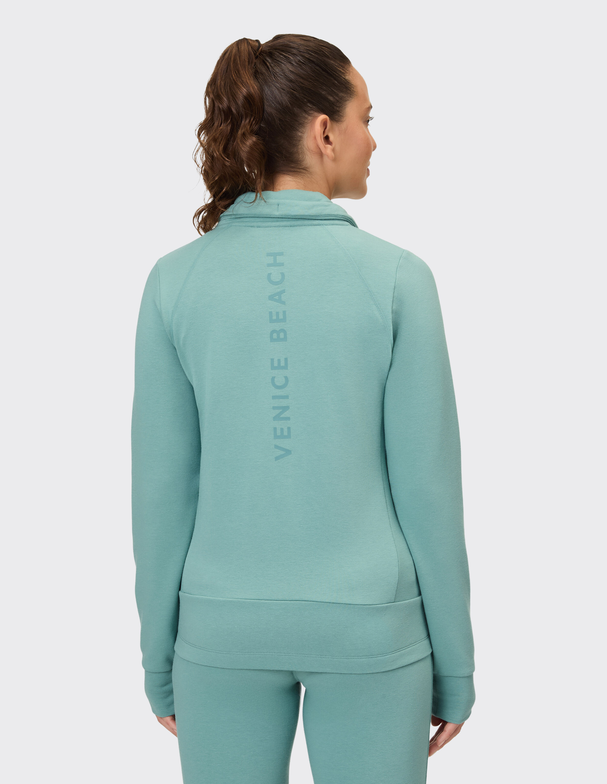 Frau trägt eine gemütliche mintgrüne Freizeitjacke mit Reißverschluss und Venice Beach-Aufdruck auf der Rückseite.