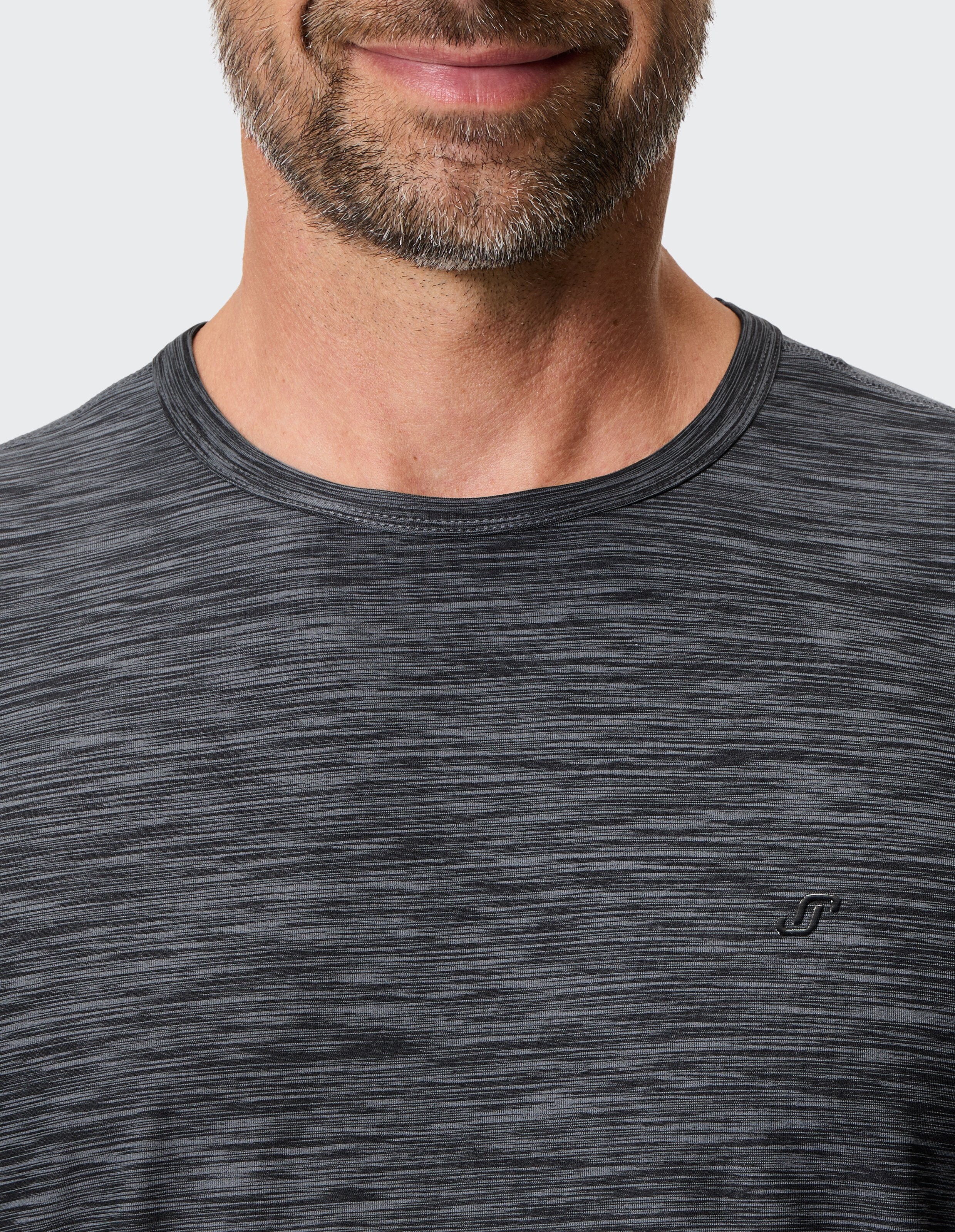 Ein Mann mit Bart trägt ein grau meliertes T-Shirt mit kleinem Logo auf der Brust, sein Gesicht zeigt ein leichtes Lächeln.