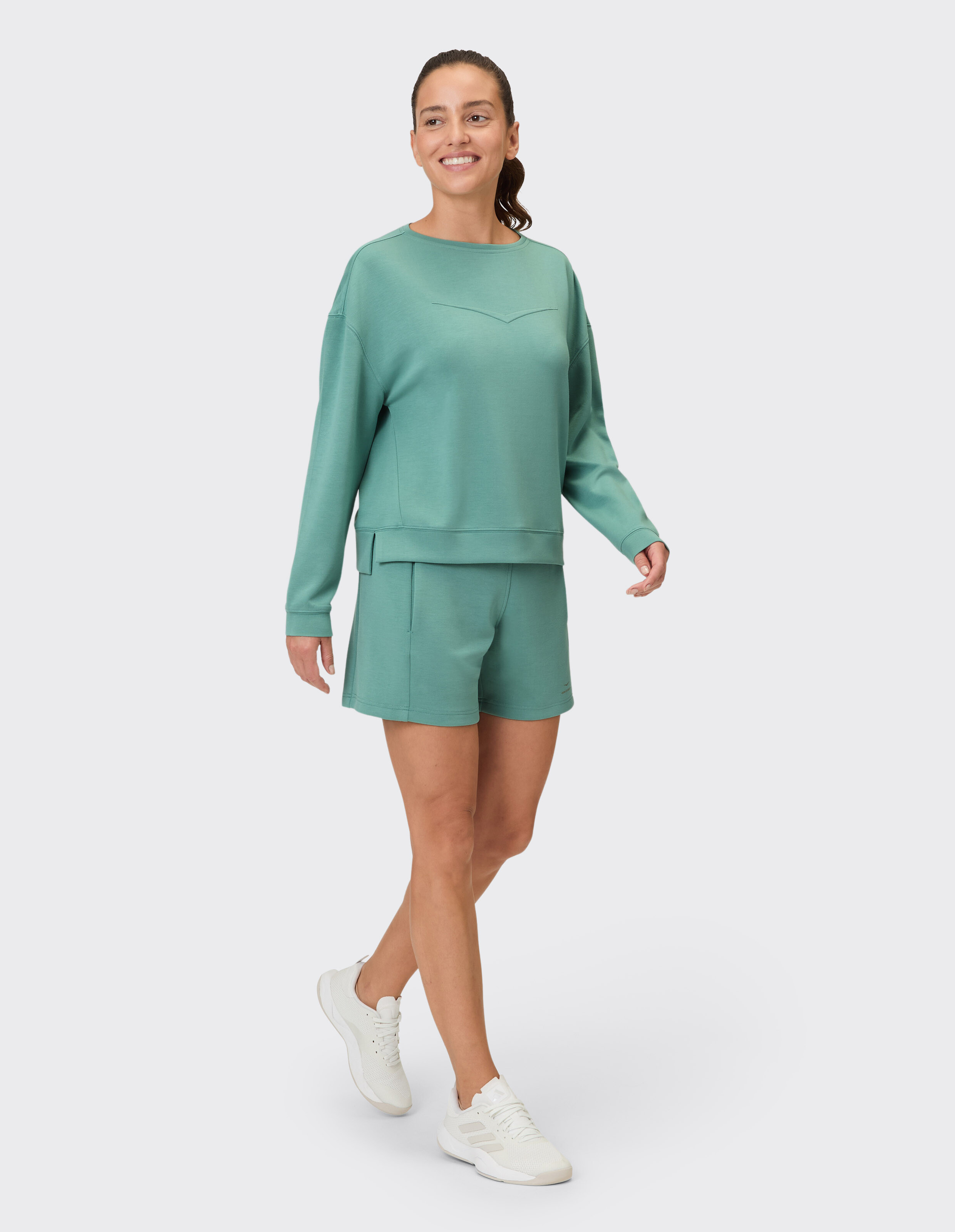 Frau trägt ein gemütliches salbeigrünes Lounge-Sweatshirt und passende lässige Shorts.