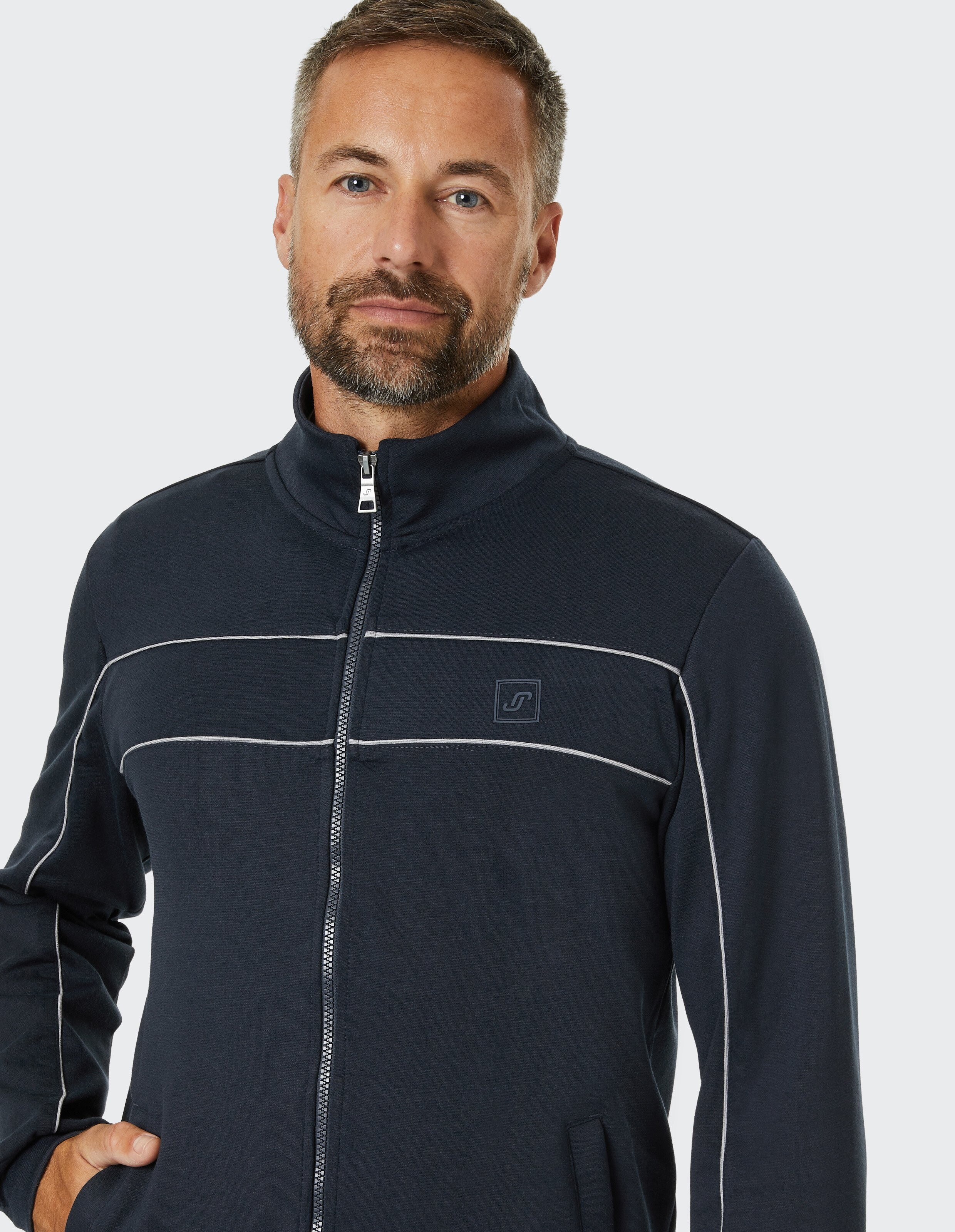 Ein Mann mit braunem Haar und Bart trägt eine dunkelblaue Jacke mit hellem Reißverschluss und dezenten, hellen Streifen.
