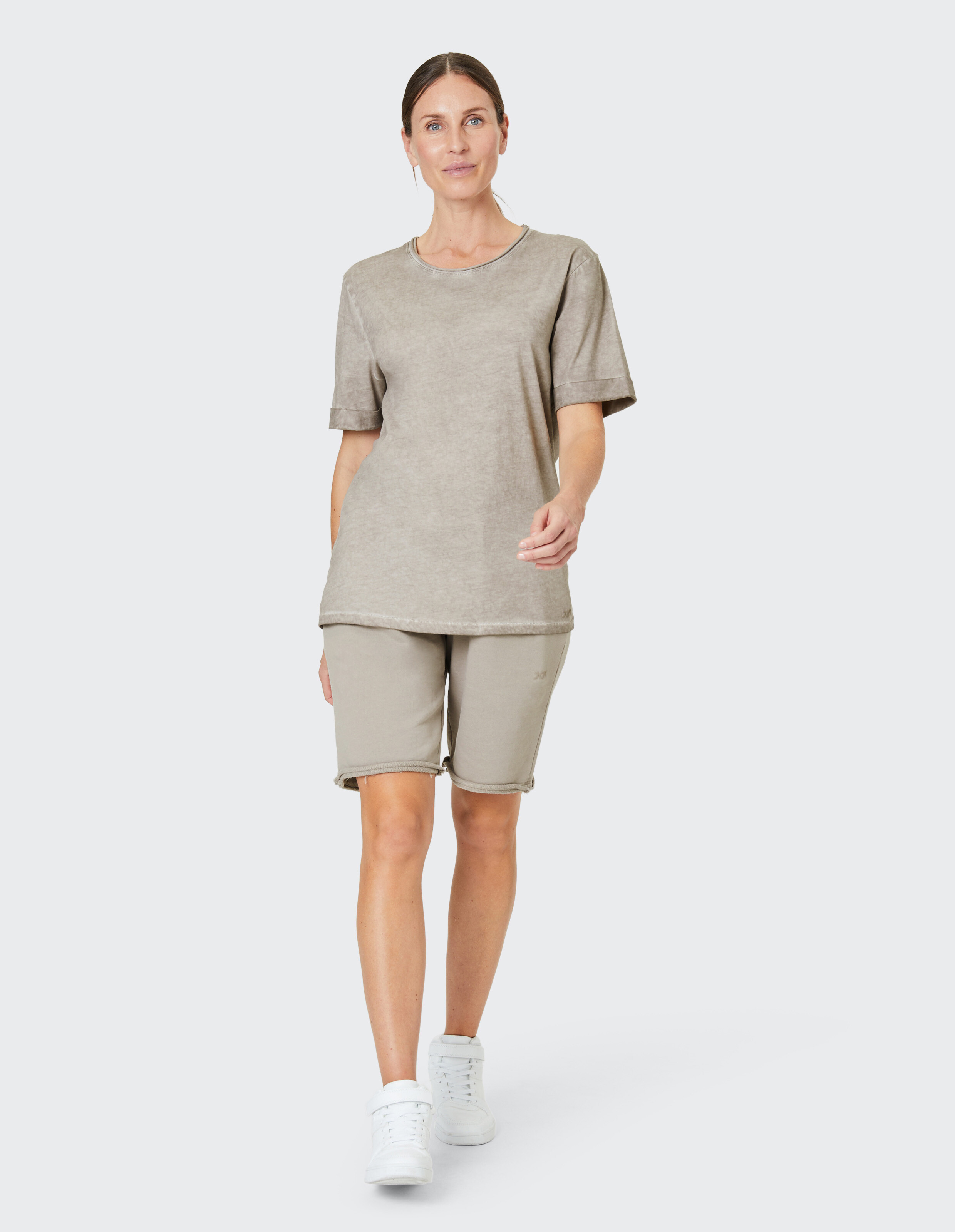 Frau in beigefarbenem T-Shirt und Shorts lächelt leicht, während sie in weißen Sneakers auf neutralem Hintergrund läuft.