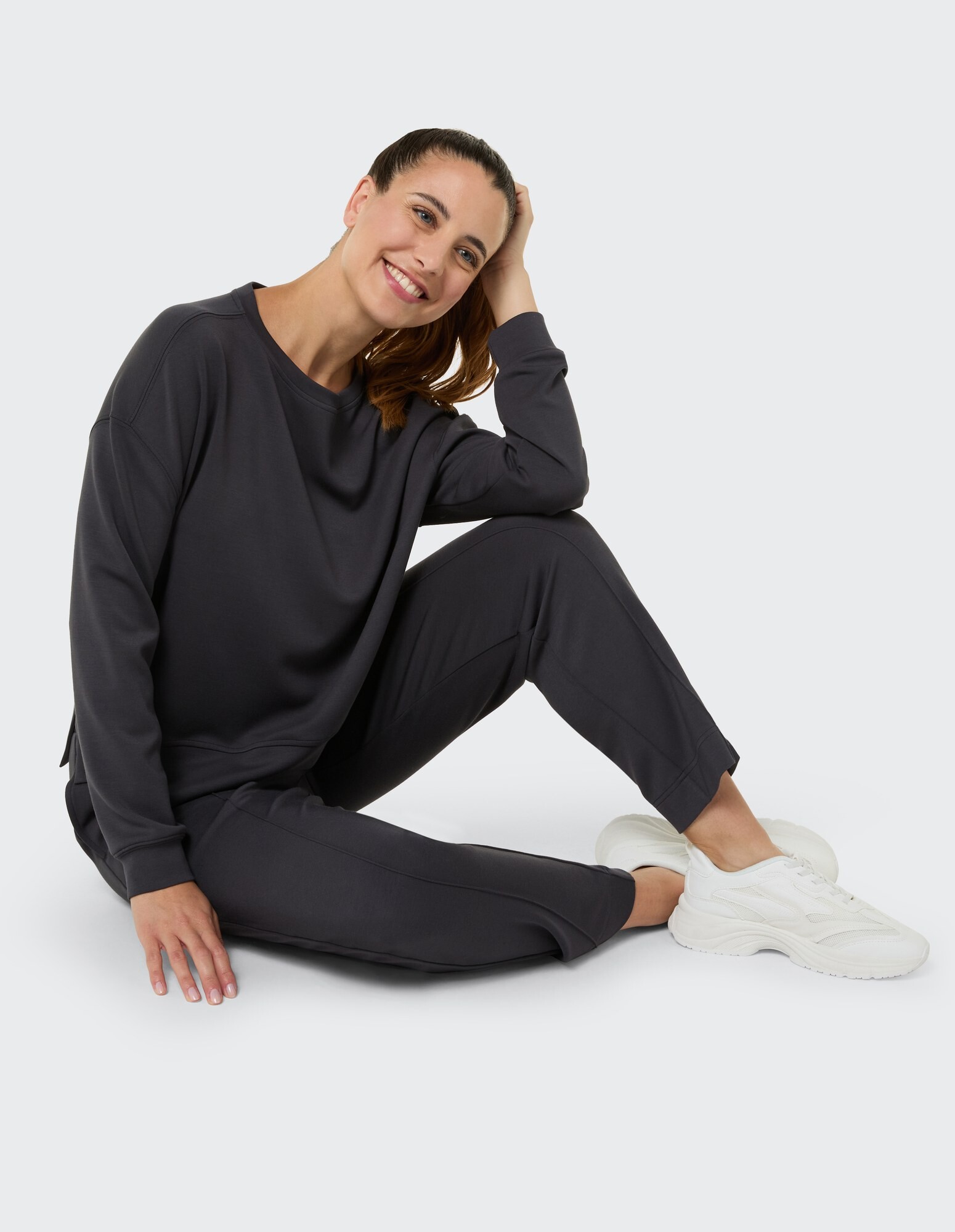 Lächelnde Frau in gemütlichem dunkelgrauem Loungewear-Set, lässig sitzend, mit weißen Turnschuhen.