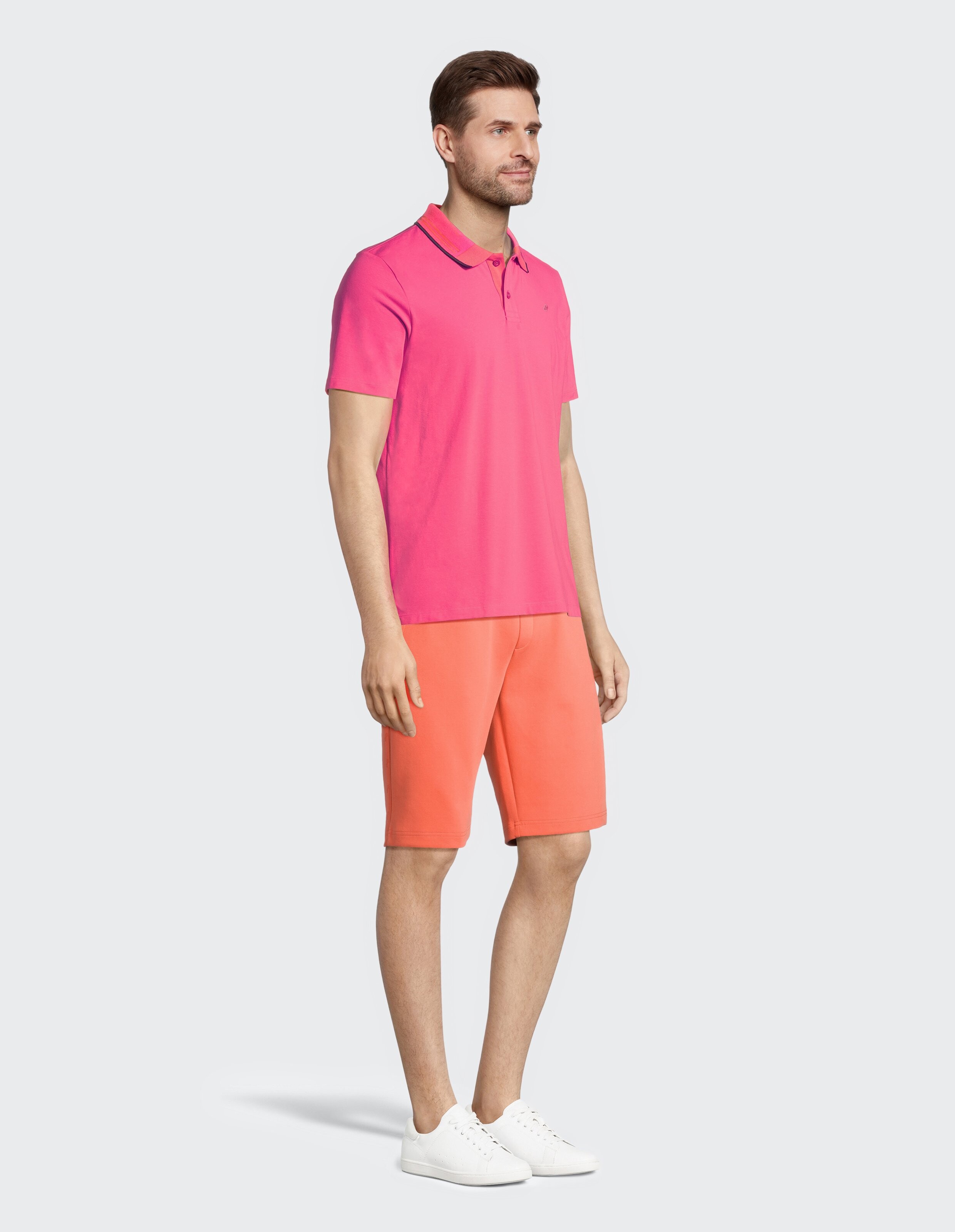 Ein Mann trägt ein pinkes Polohemd, orange Shorts und weiße Sneakers vor einem weißen Hintergrund, mit neutralem Lächeln.