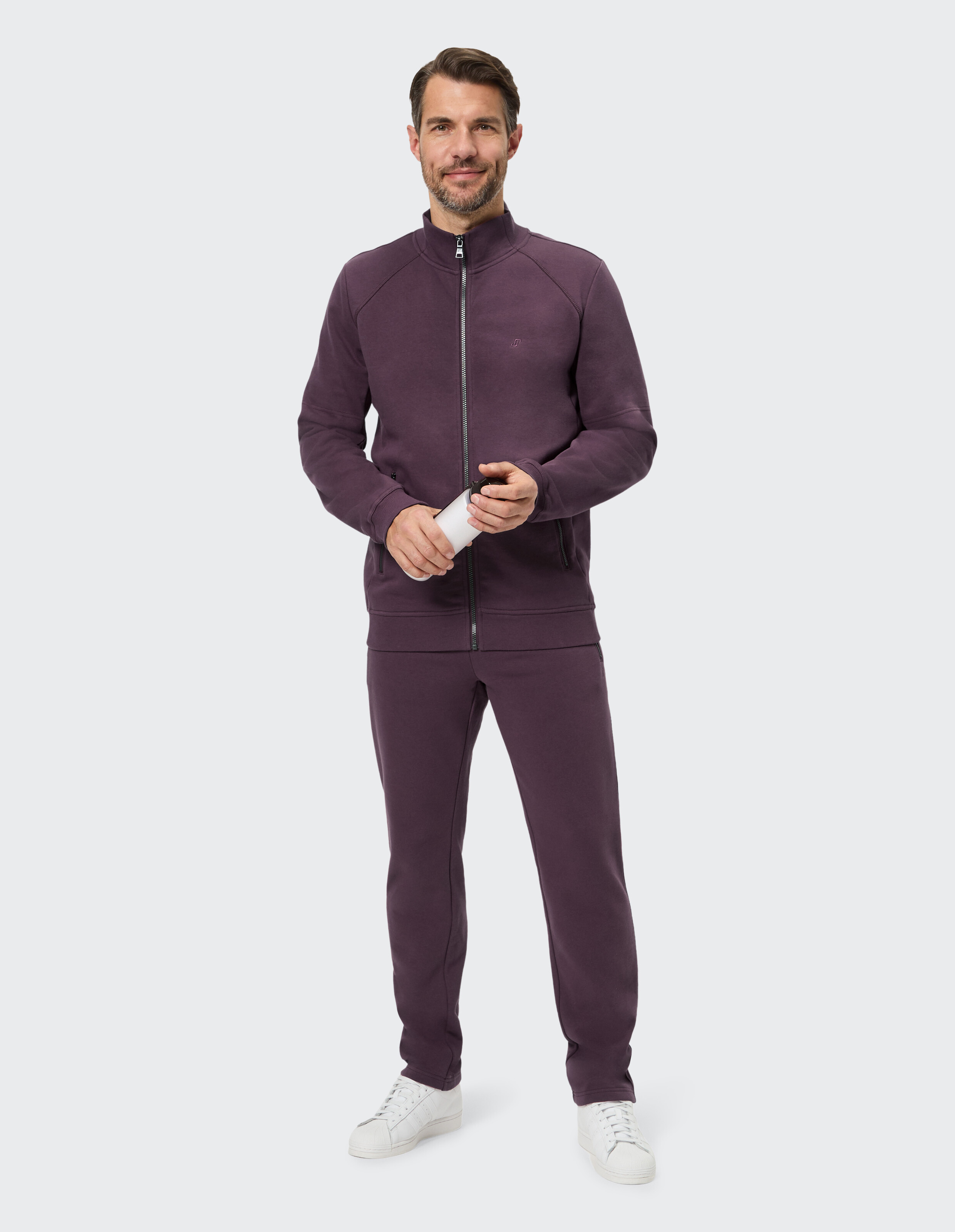 Mann in lilafarbener Jacke mit Reißverschluss und passender Hose, hält ein Getränk in der Hand.