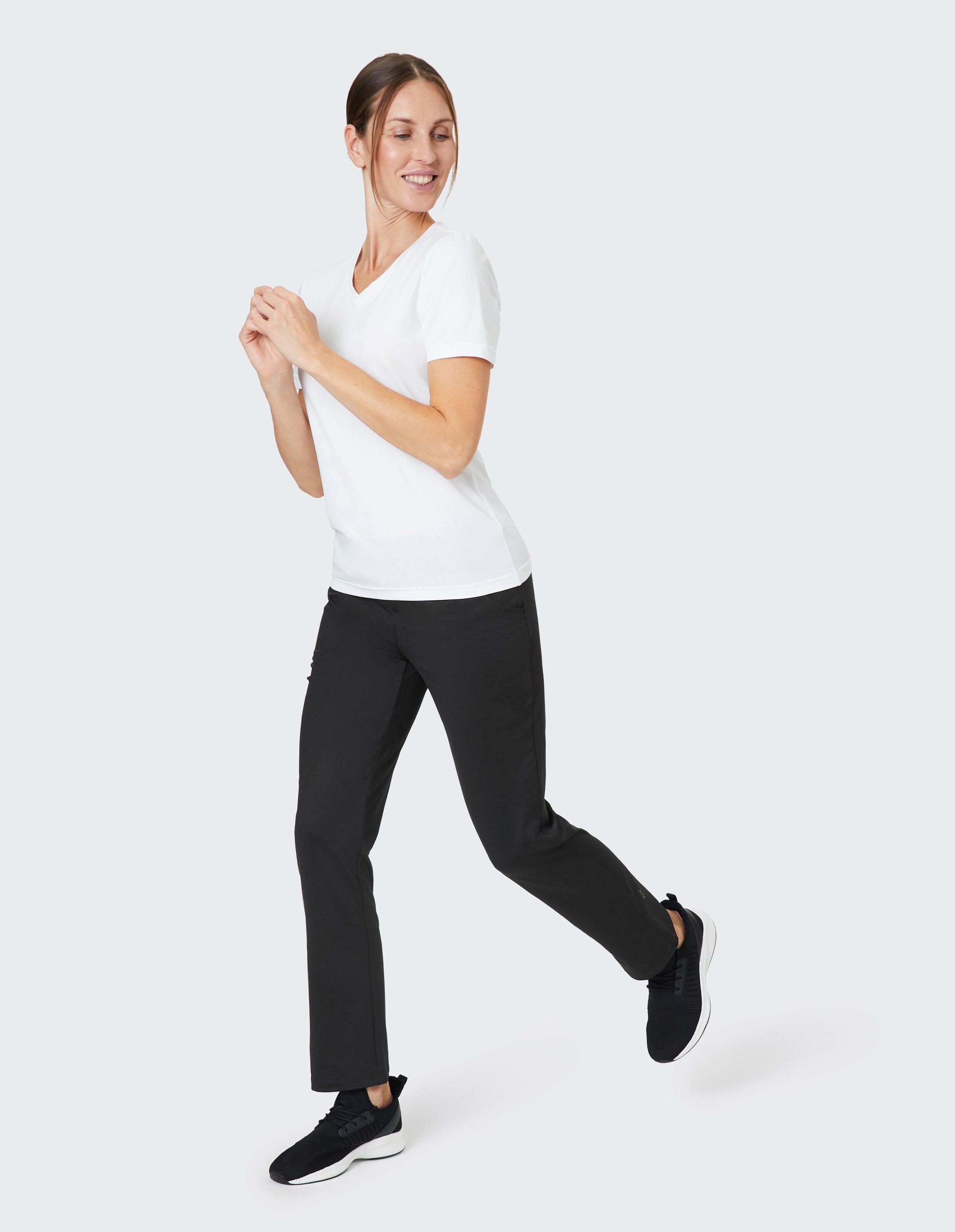 Frau in weißem T-Shirt und schwarzer Hose lächelt und macht einen Laufbewegung vor grauem Hintergrund, trägt schwarze Turnschuhe.