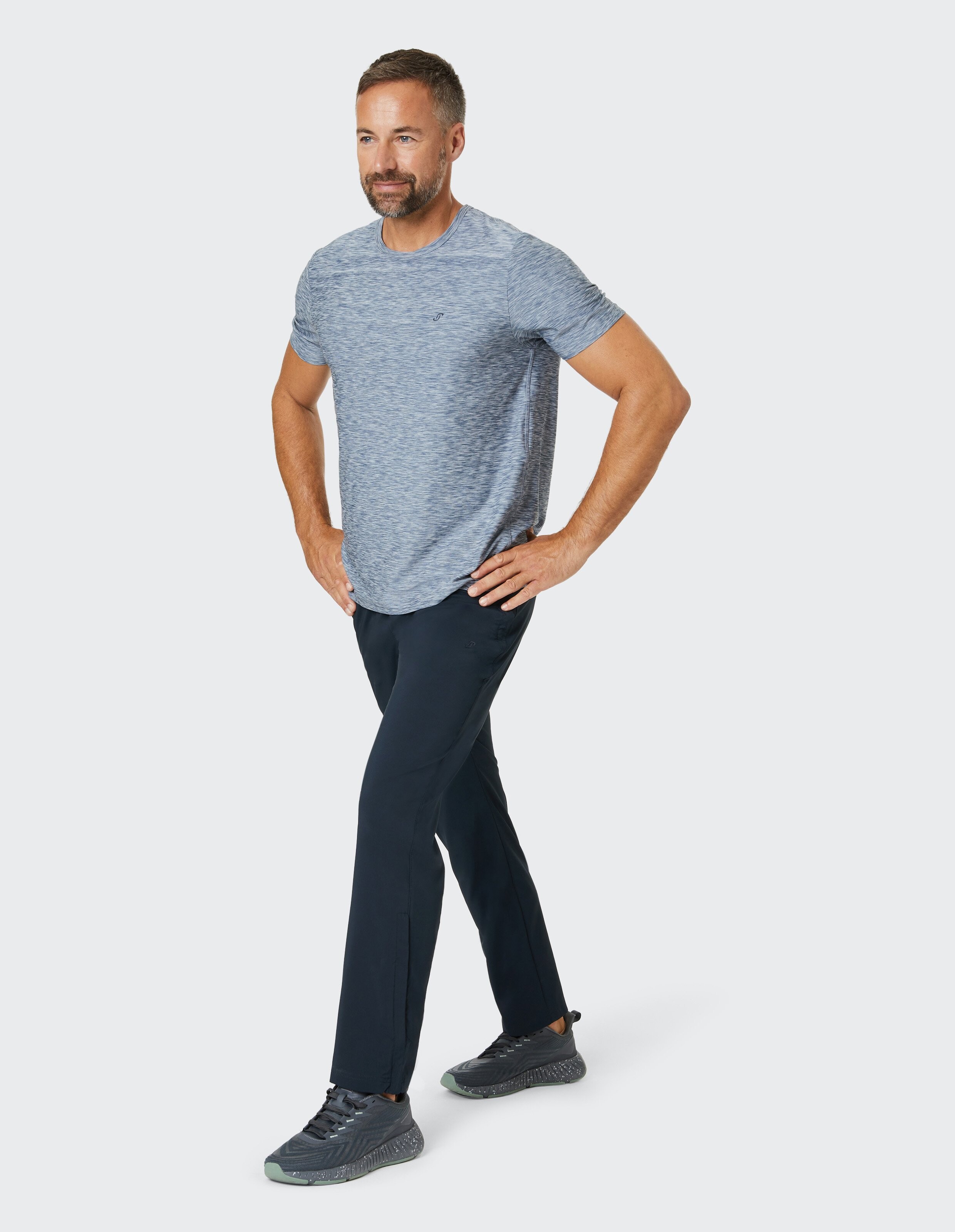 Ein Mann in grauem T-Shirt und dunkler Hose steht entspannt mit den Händen in den Hüften, blickt nach vorn und trägt Sportschuhe.