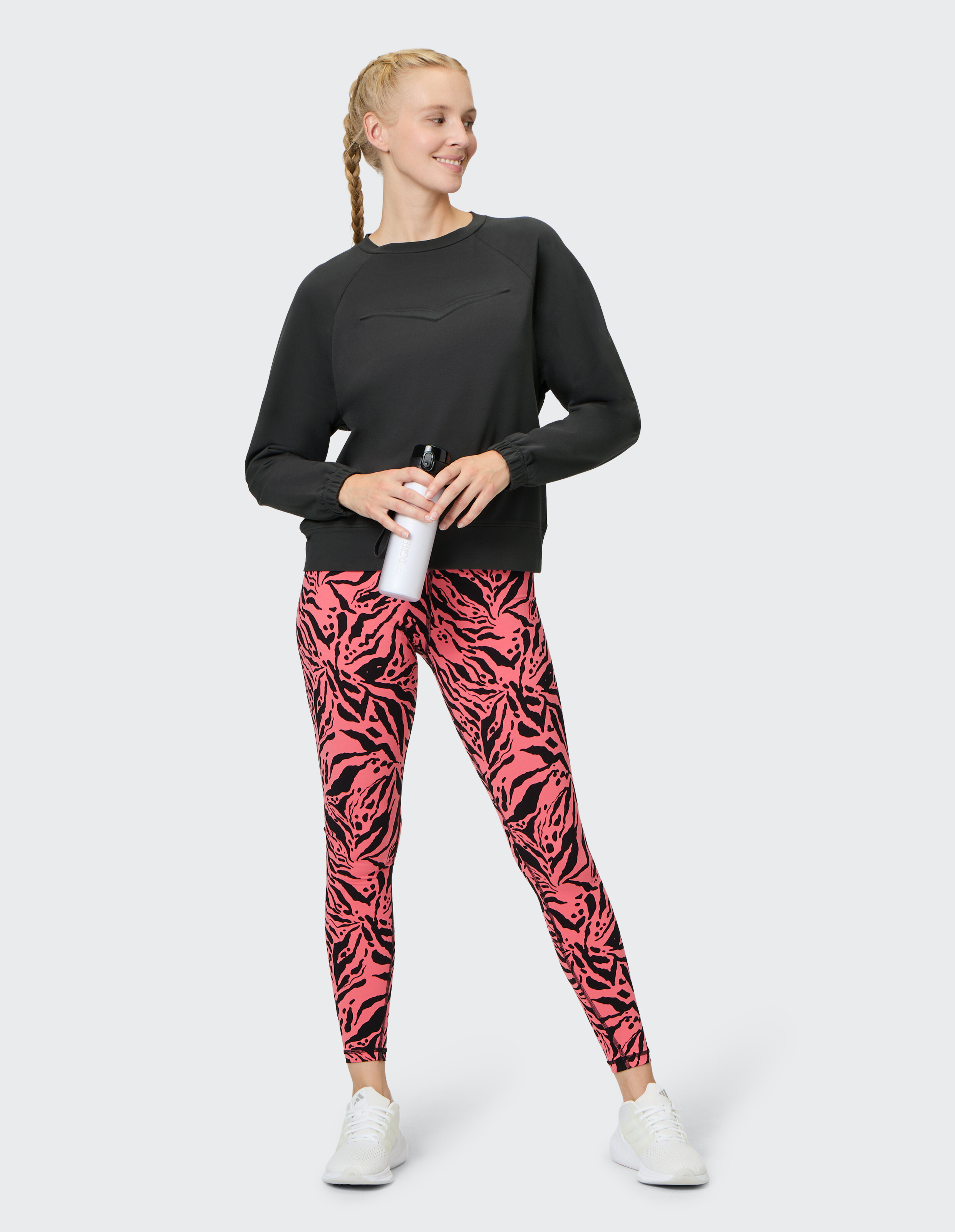 Frau in schwarzem Sweatshirt und rosa gemusterten Leggings, hält eine Wasserflasche in der Hand.