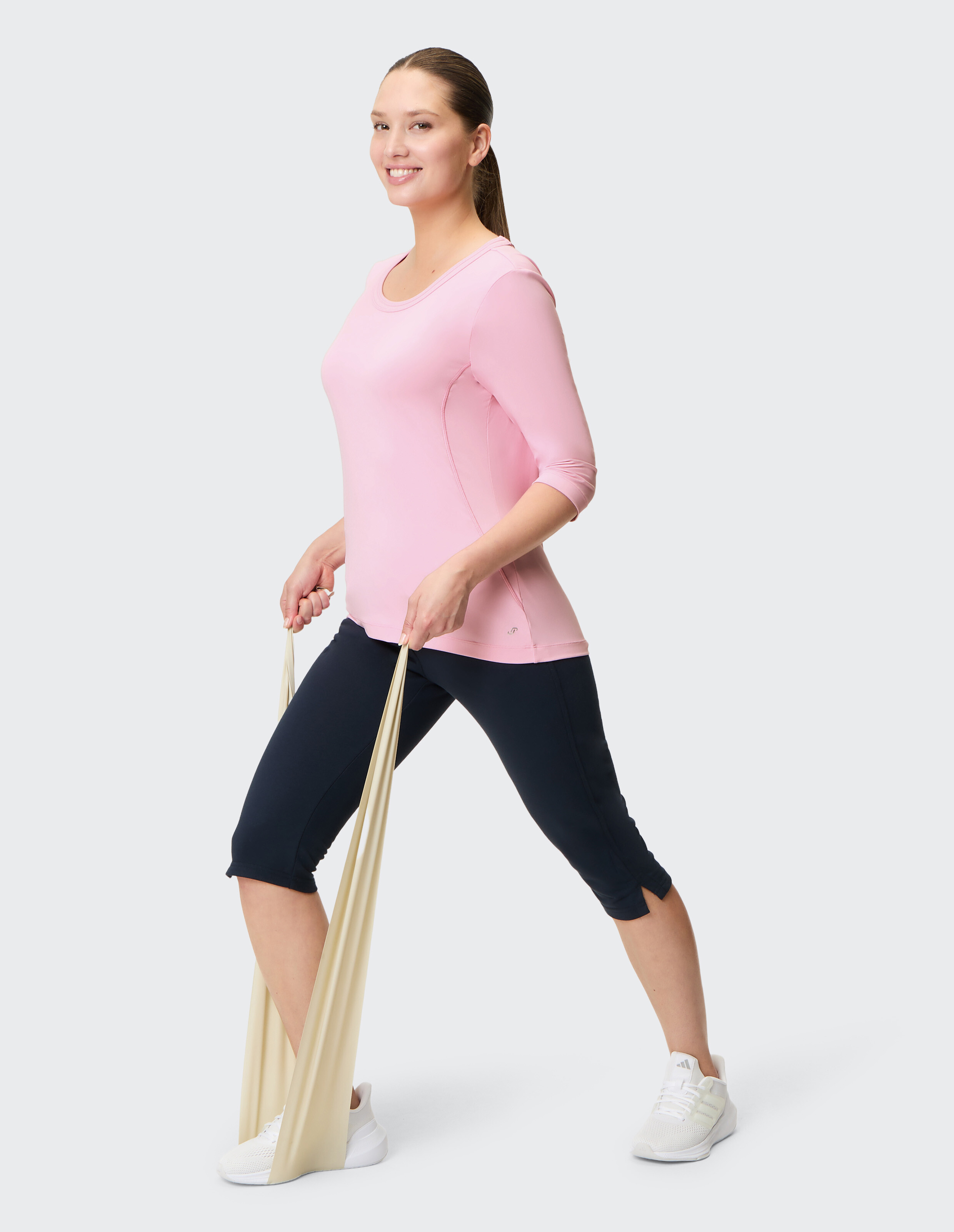 Frau in rosa Shirt und schwarzen Leggings trainiert mit einem beige-farbenen Widerstandsband, während sie lächelnd nach vorne schaut.